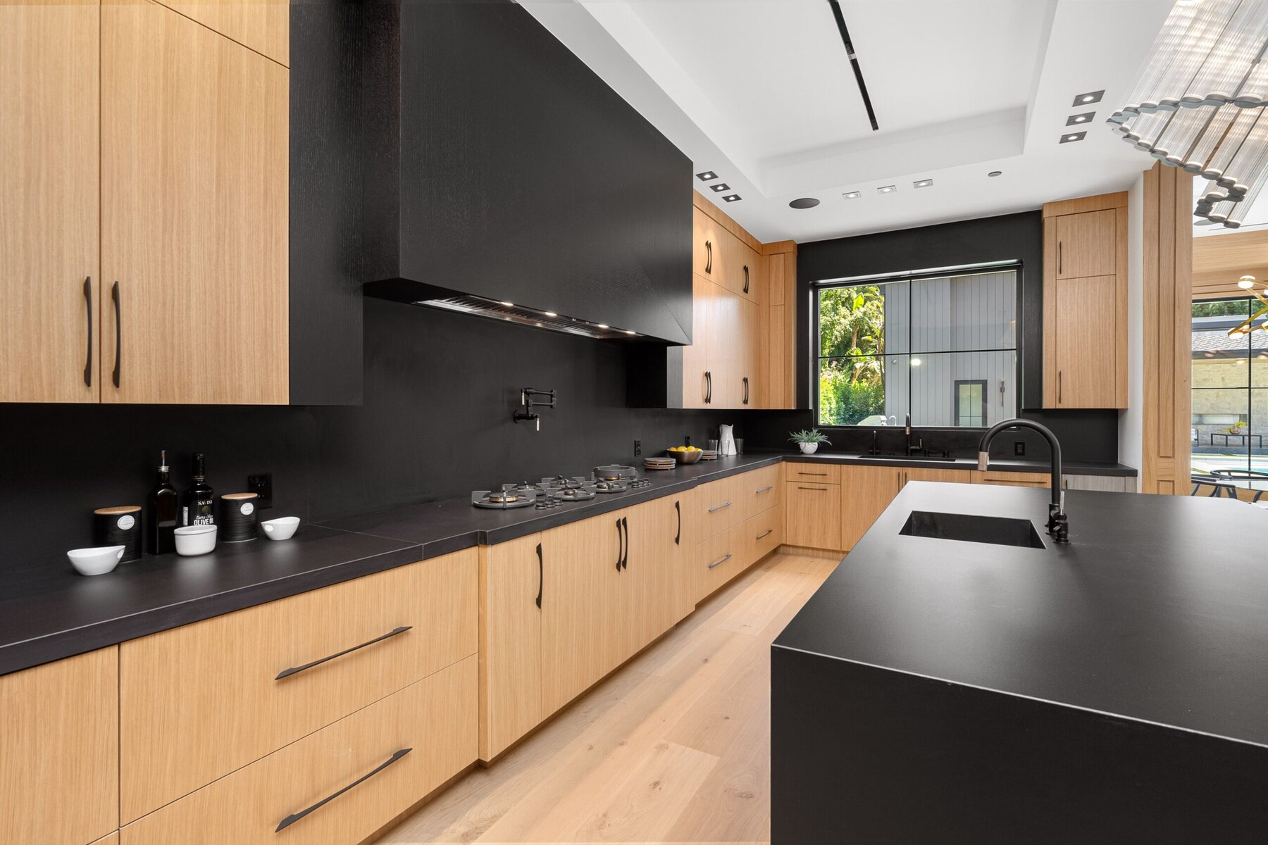 Modern kitchen with light wood cabinets, black countertops, and a large window.