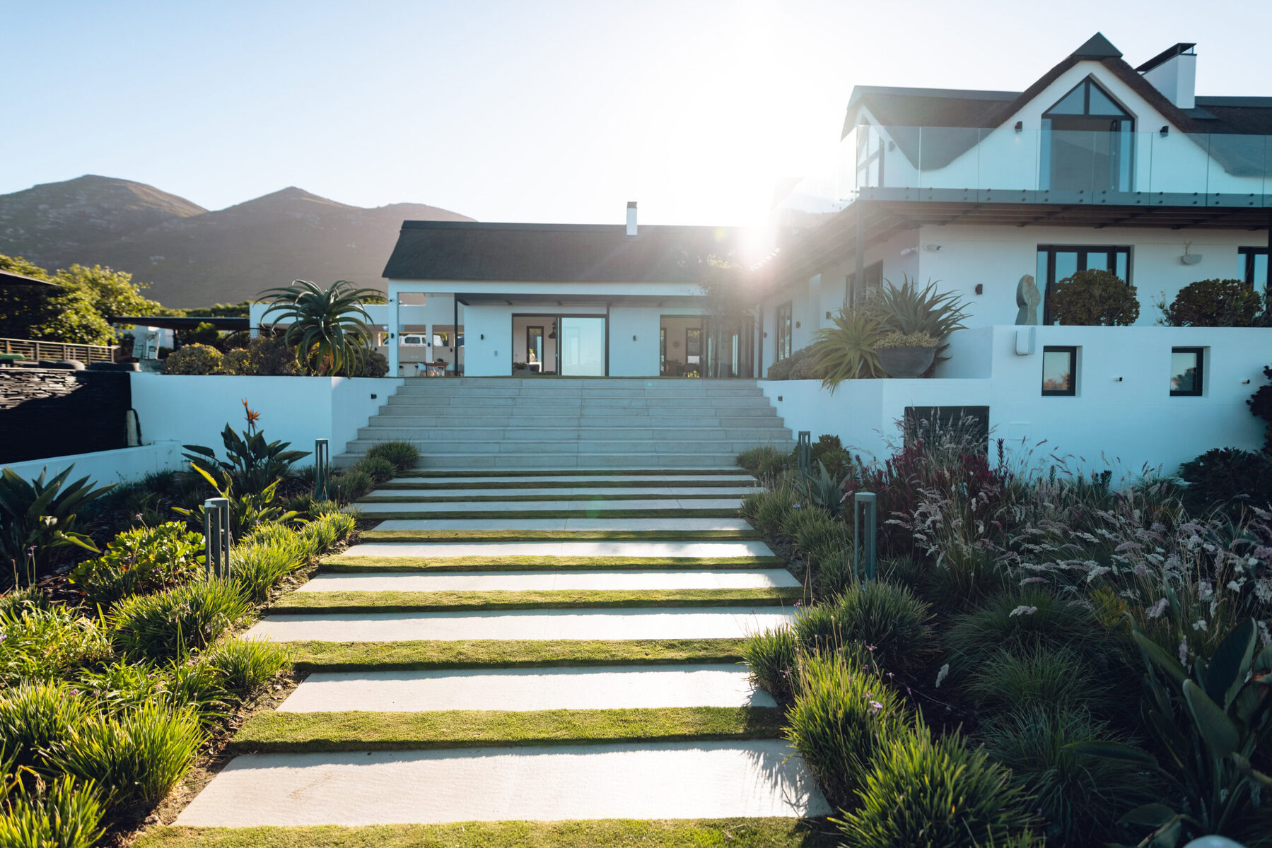 Modern white house with terraced garden and mountain backdrop under bright sun.