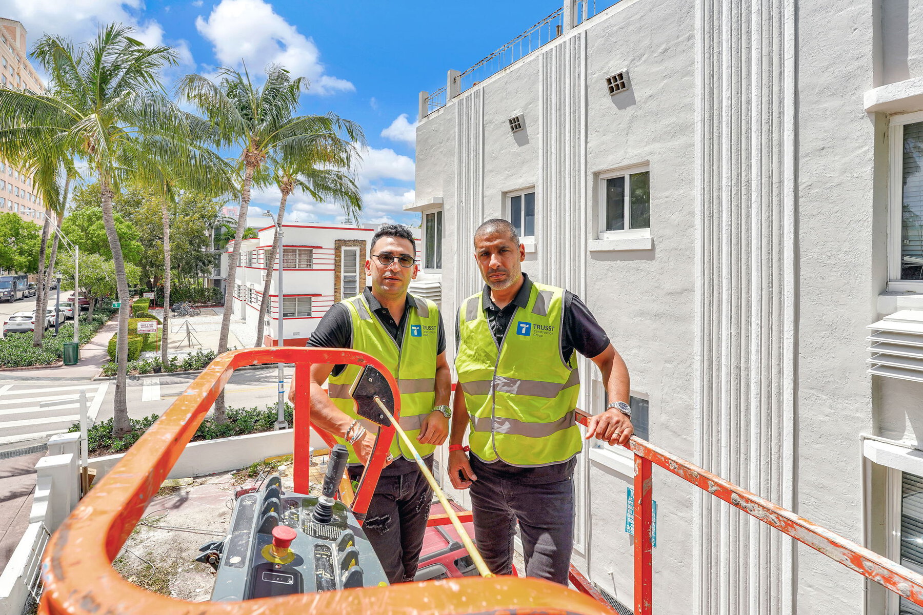 Two men in safety vests on a lift next to a white building.