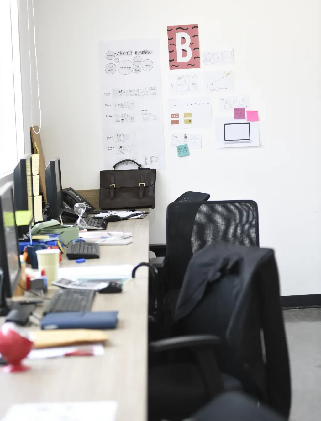 Messy office desk with a briefcase, monitors, and papers on the wall.