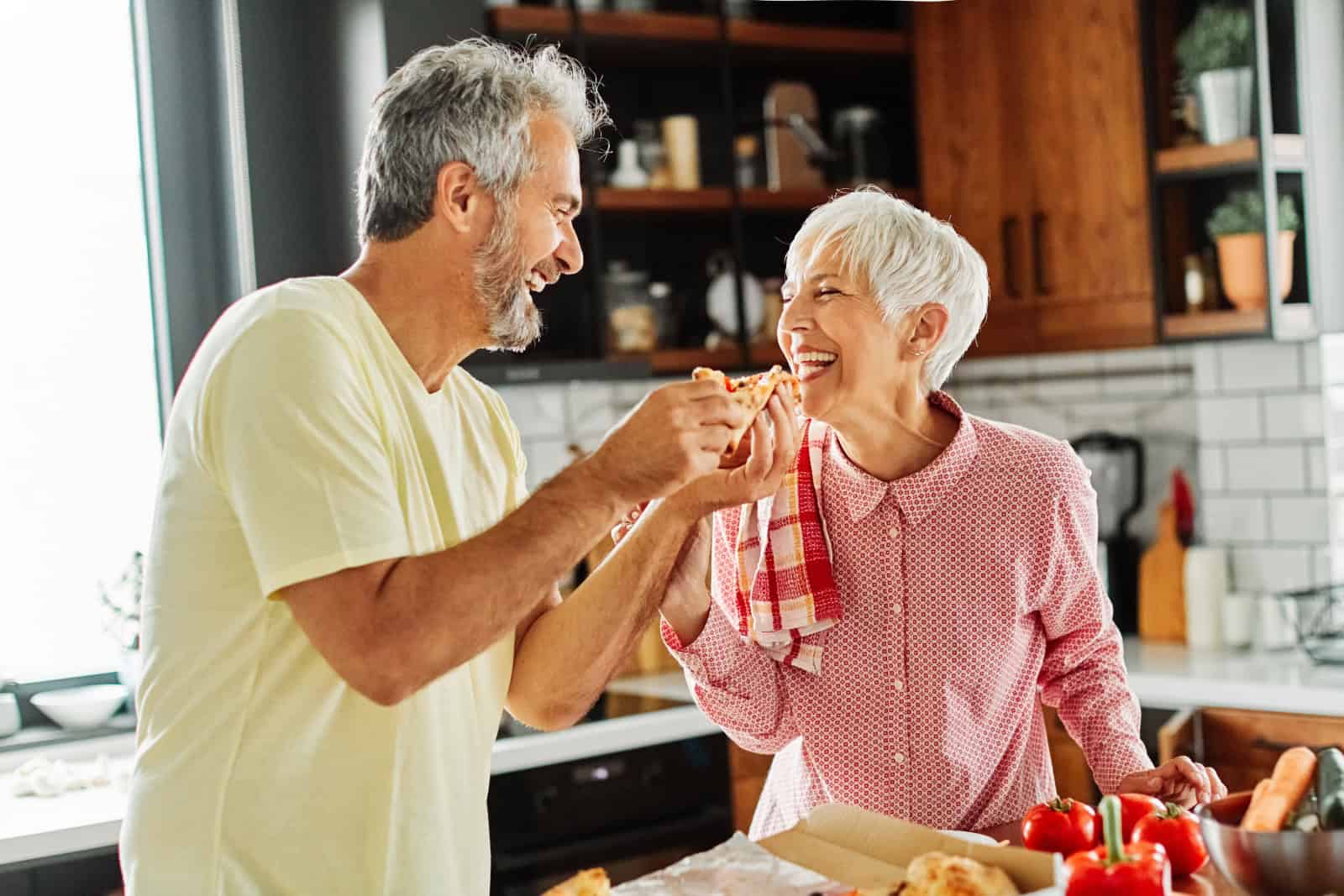 Portrait of happy senior couple prepering meal and tasting pizza in kitchen