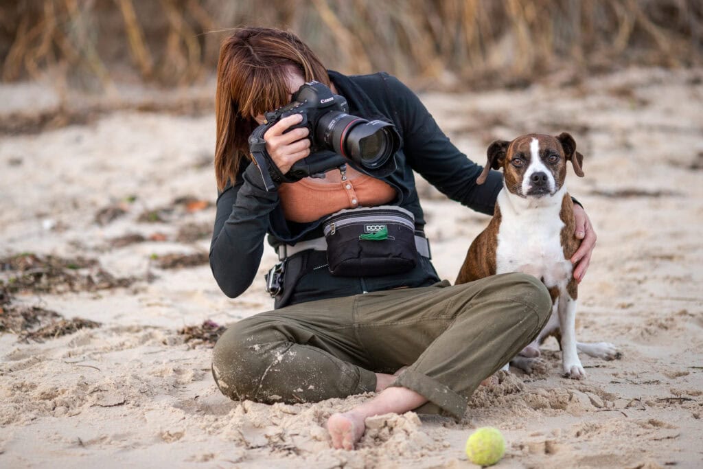 a photographer with a camera in hand and dog on her left photographing another dog 