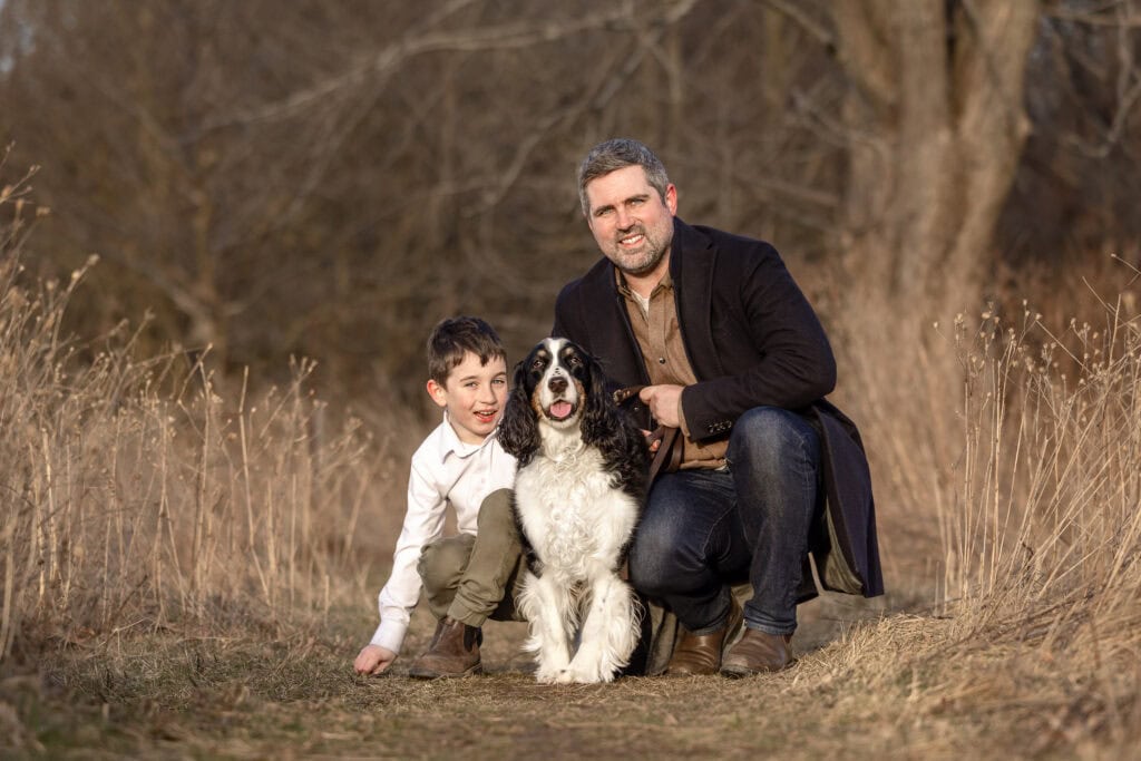 A man and a boy with a dog sitting in  grass in the Spring at Niagara Shores park in Niagara on the Lake