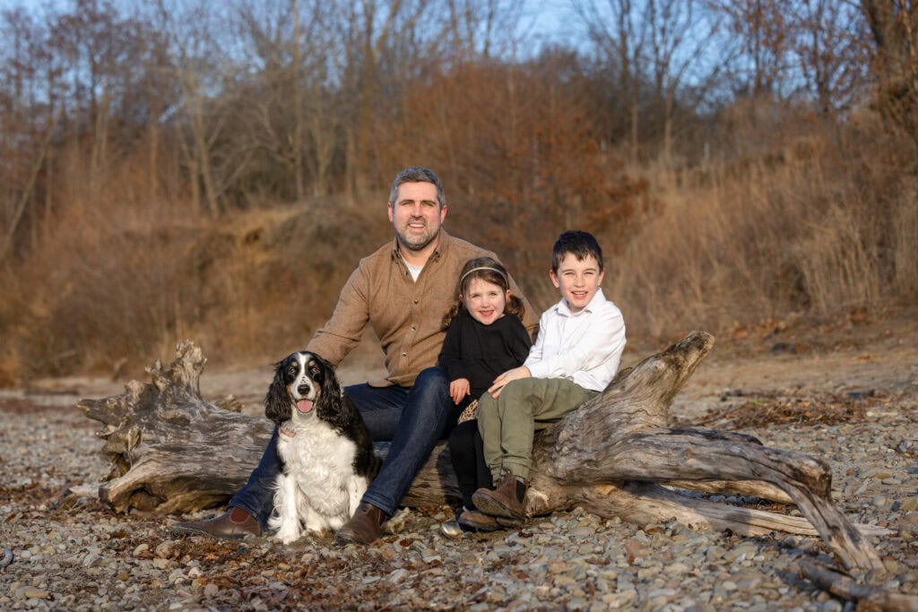 A man and two children giggling with their dog sitting on a log at Niagara Shores Park in Niagara on the Lake