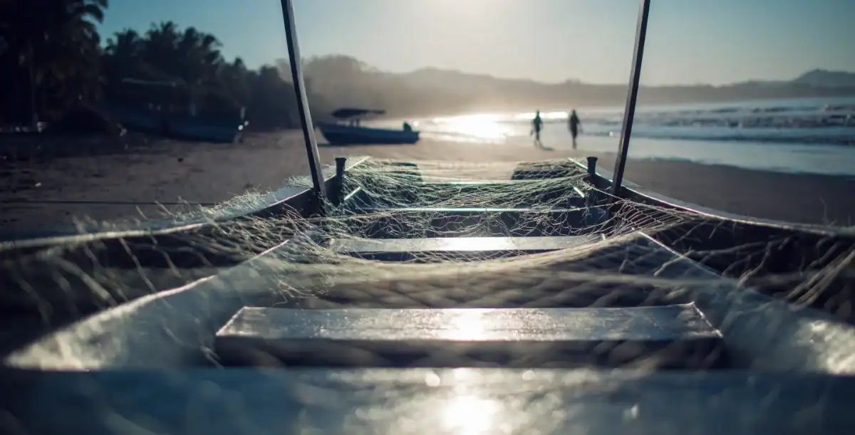Vista desde la orilla de una playa con una red de pesca y remos, con personas y barcos en el fondo durante el atardecer.