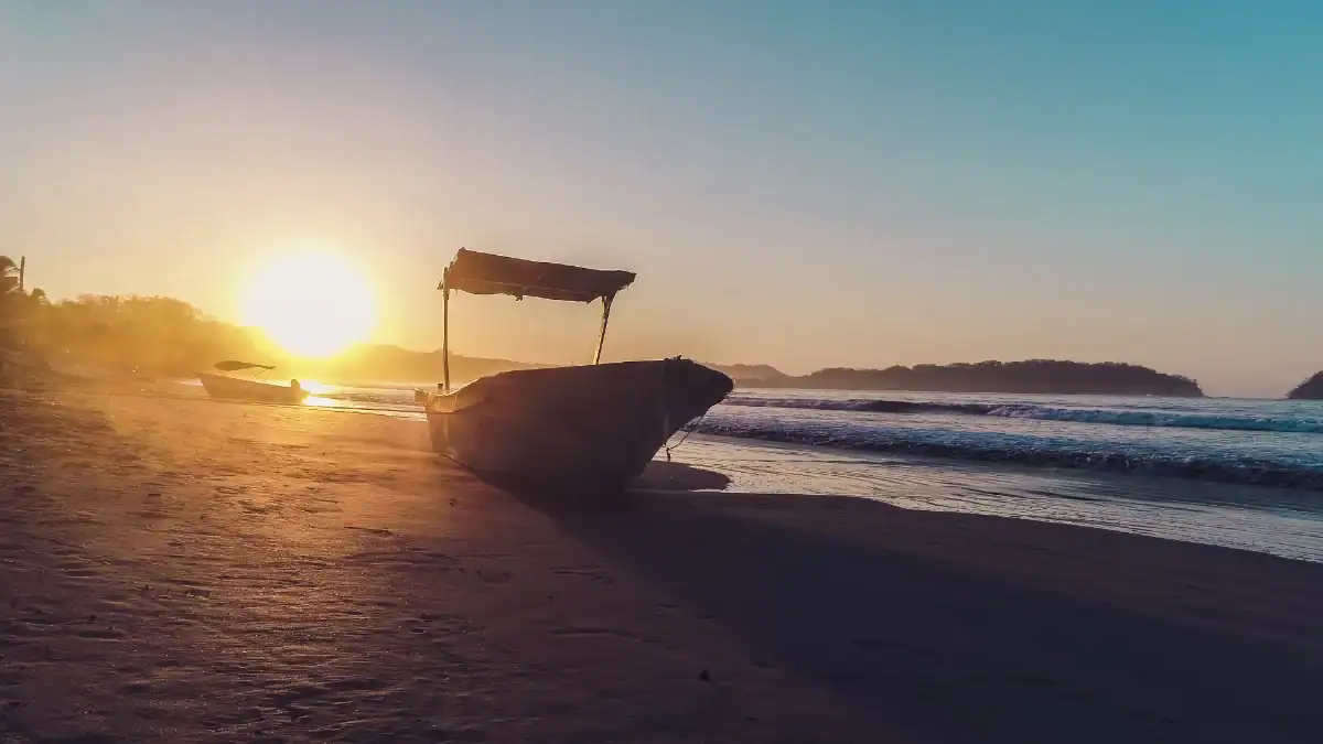 Barco en la playa durante el atardecer con el sol en el horizonte y olas suaves en el mar.