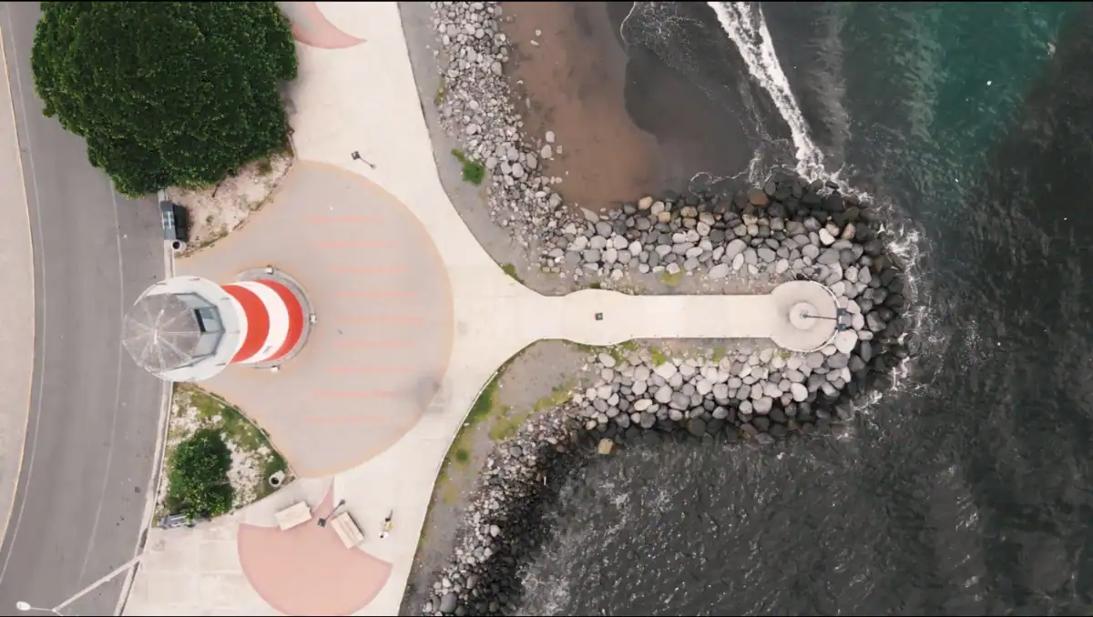 Vista aérea de un faro en un muelle de piedra en la costa, con olas rompiendo contra las rocas y un camino que lleva al faro.