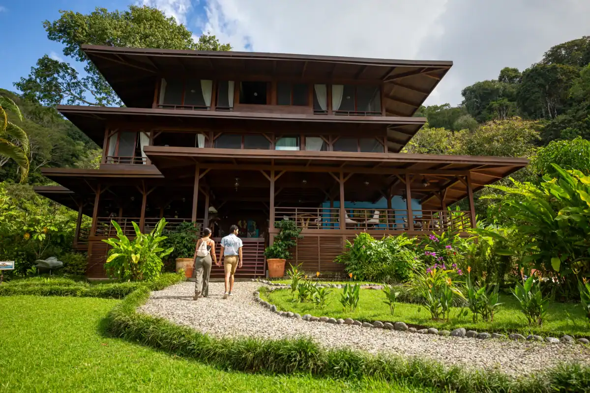 Casa de madera de tres pisos en un entorno natural con jardín y sendero de grava, dos personas caminando hacia la entrada.