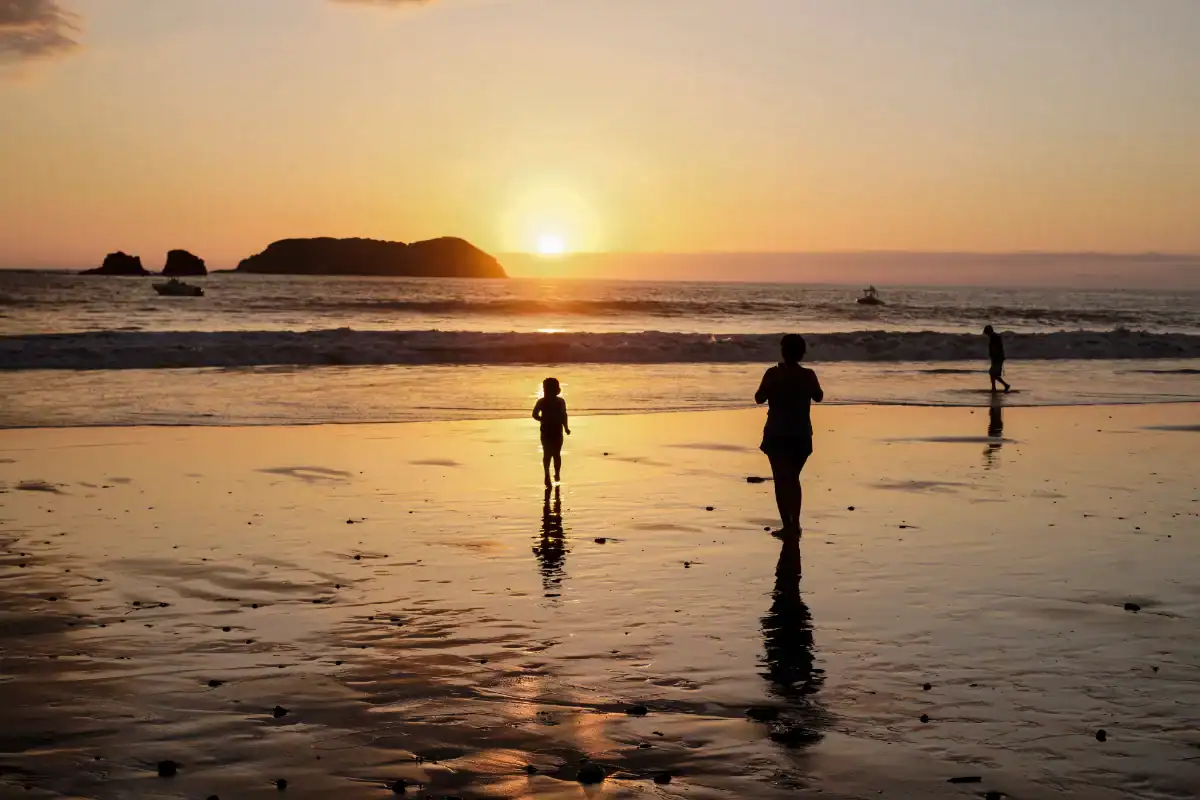 Personas caminando en la playa durante la puesta del sol, con un fondo de mar y una isla en el horizonte.
