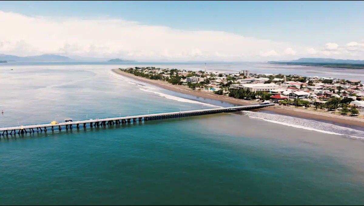 Vista aérea de un muelle que se extiende hacia el mar, con una ciudad costera y montañas al fondo.