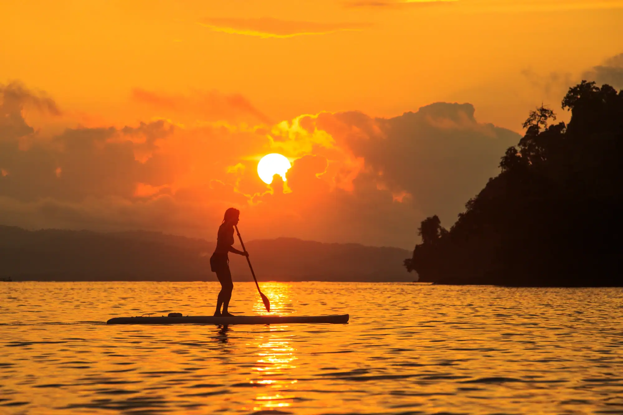 Persona haciendo paddle surf en un lago al atardecer con cielo naranja y nubes.