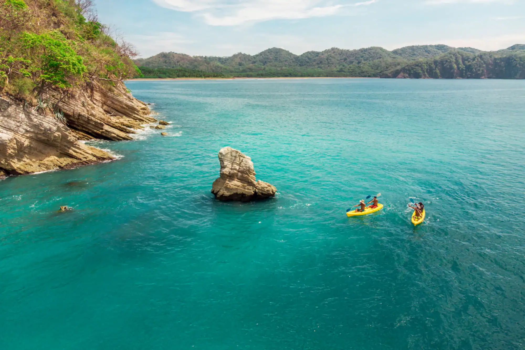 Dos personas en kayak en aguas turquesas cerca de una costa con vegetaci├│n y monta├▒as al fondo.