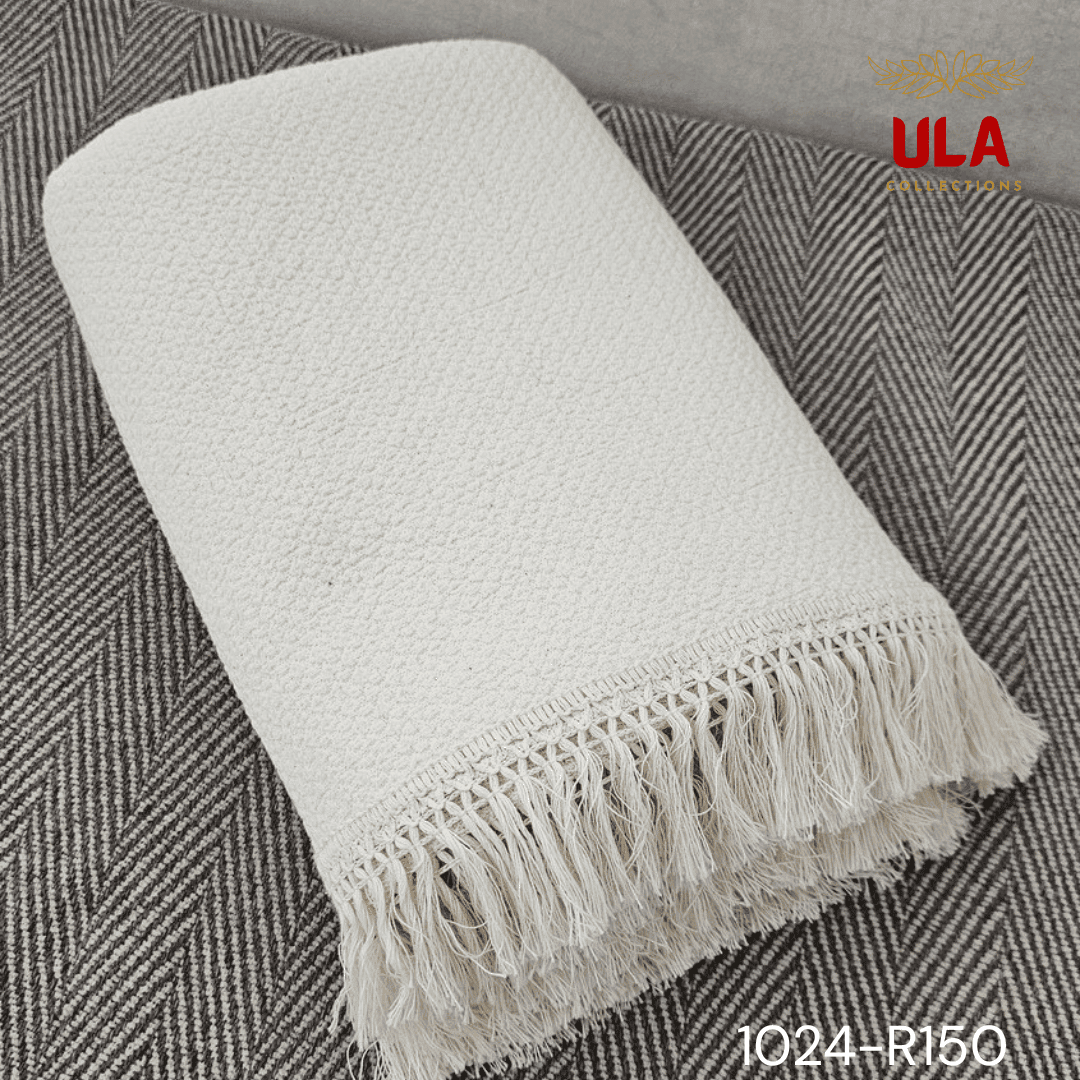 A folded cream or off-white woven throw blanket with a prominent basketweave or waffle knit texture. The textile is finished with a wide natural-colored tassel fringe and is displayed draped over a couch or background fabric with a dark gray herringbone pattern, highlighting a modern farmhouse or cozy minimalist home decor style.