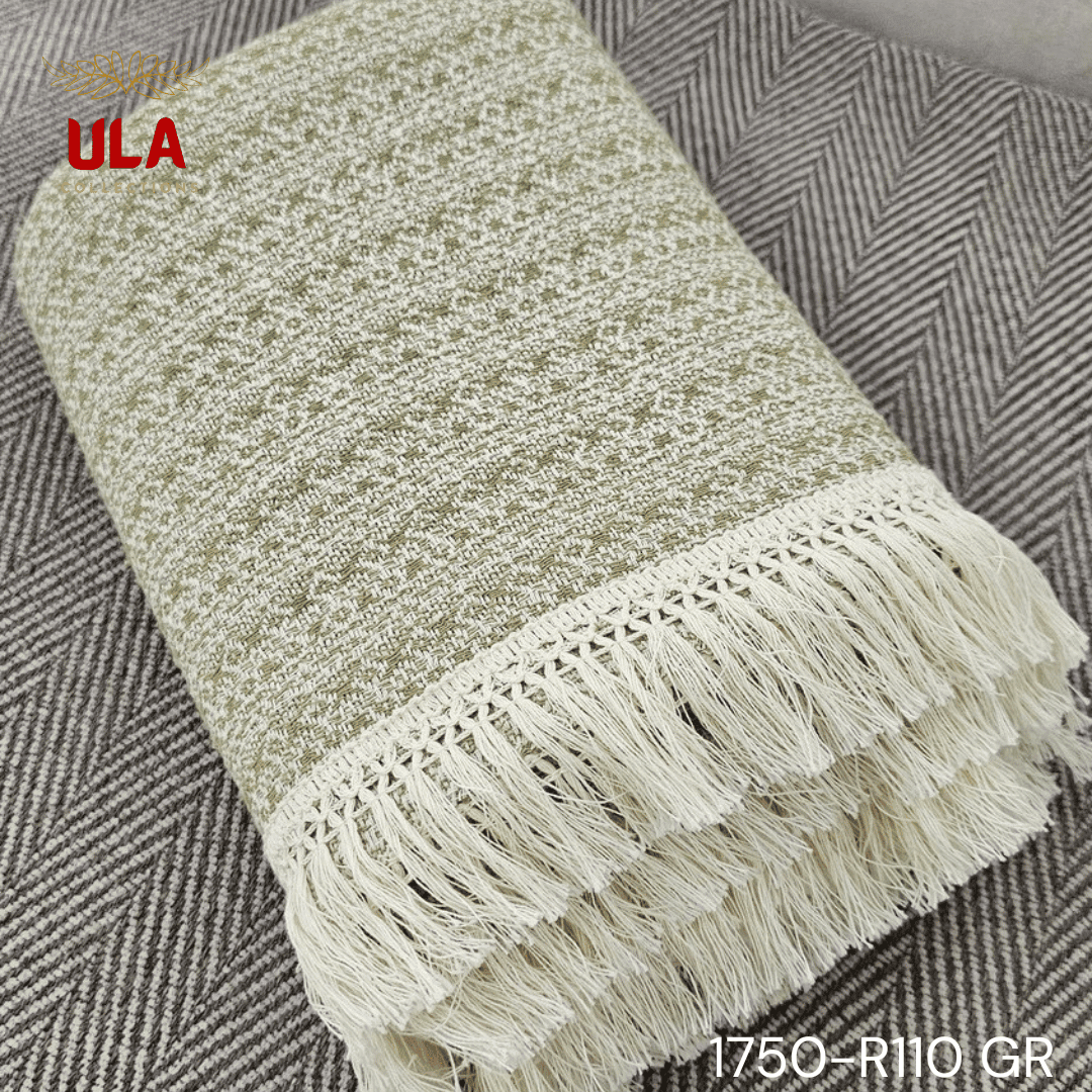 A close-up view of a soft woven throw blanket or textile, primarily in a speckled sage green or light olive color, blended with white threads for a heathered texture. The blanket has a subtle diamond or geometric pattern in its weave and is finished with a wide cream or natural-colored tassel fringe.