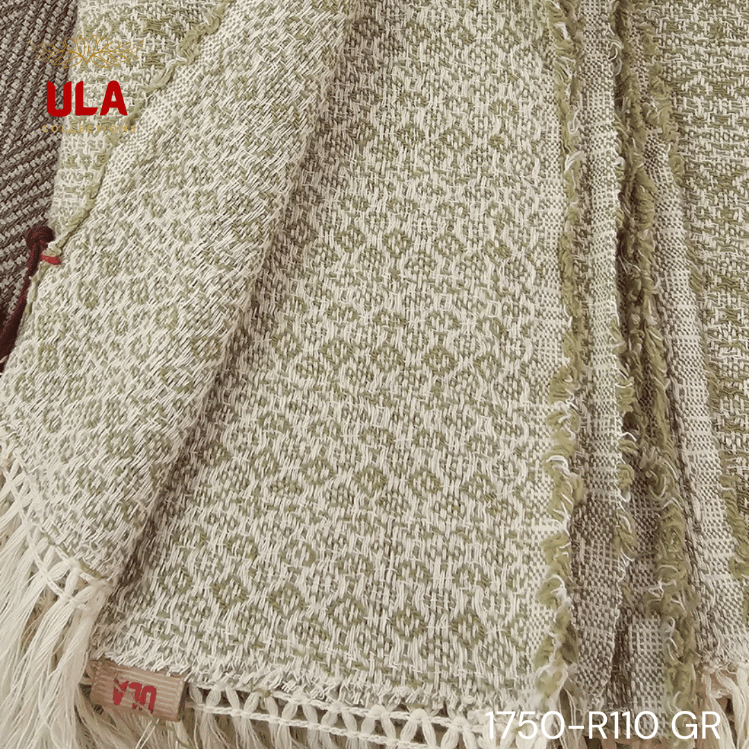 A close-up view of a soft woven throw blanket or textile, primarily in a speckled sage green or light olive color, blended with white threads for a heathered texture. The blanket has a subtle diamond or geometric pattern in its weave and is finished with a wide cream or natural-colored tassel fringe.