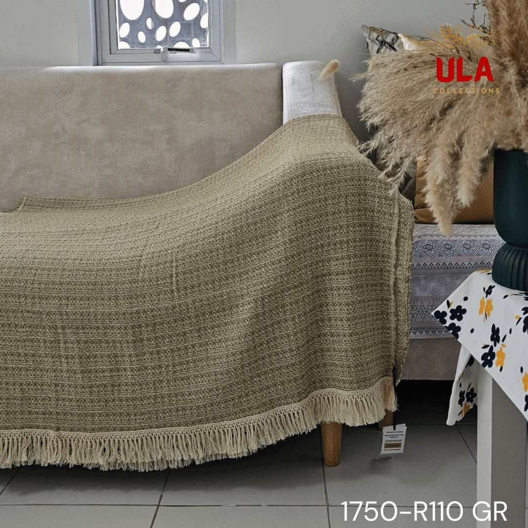 A close-up view of a soft woven throw blanket or textile, primarily in a speckled sage green or light olive color, blended with white threads for a heathered texture. The blanket has a subtle diamond or geometric pattern in its weave and is finished with a wide cream or natural-colored tassel fringe.