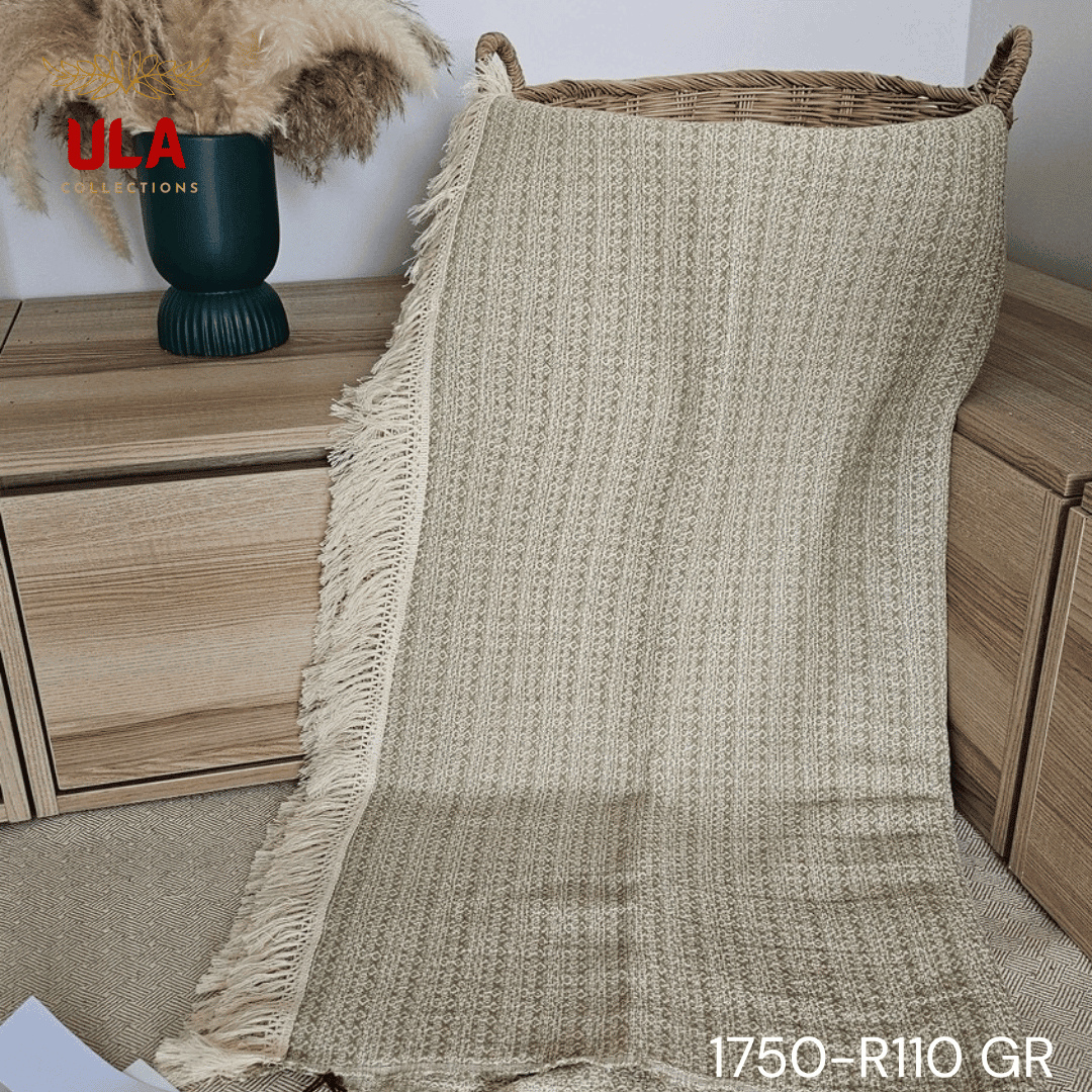 A close-up view of a soft woven throw blanket or textile, primarily in a speckled sage green or light olive color, blended with white threads for a heathered texture. The blanket has a subtle diamond or geometric pattern in its weave and is finished with a wide cream or natural-colored tassel fringe.