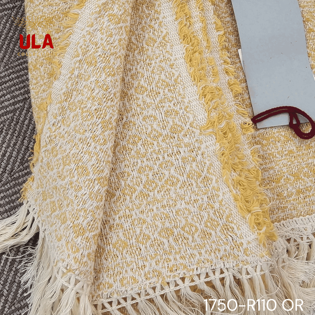A close-up view of a soft woven throw blanket or textile, primarily in a speckled mustard yellow or marigold color, blended with white threads for a heathered texture. The blanket is finished with a wide white tassel fringe