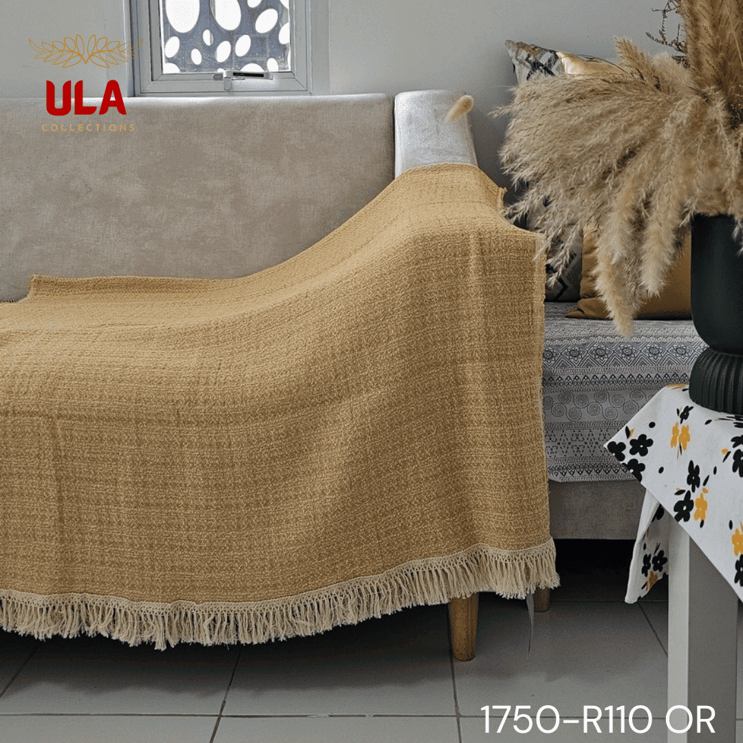 A close-up view of a soft woven throw blanket or textile, primarily in a speckled mustard yellow or marigold color, blended with white threads for a heathered texture. The blanket is finished with a wide white tassel fringe