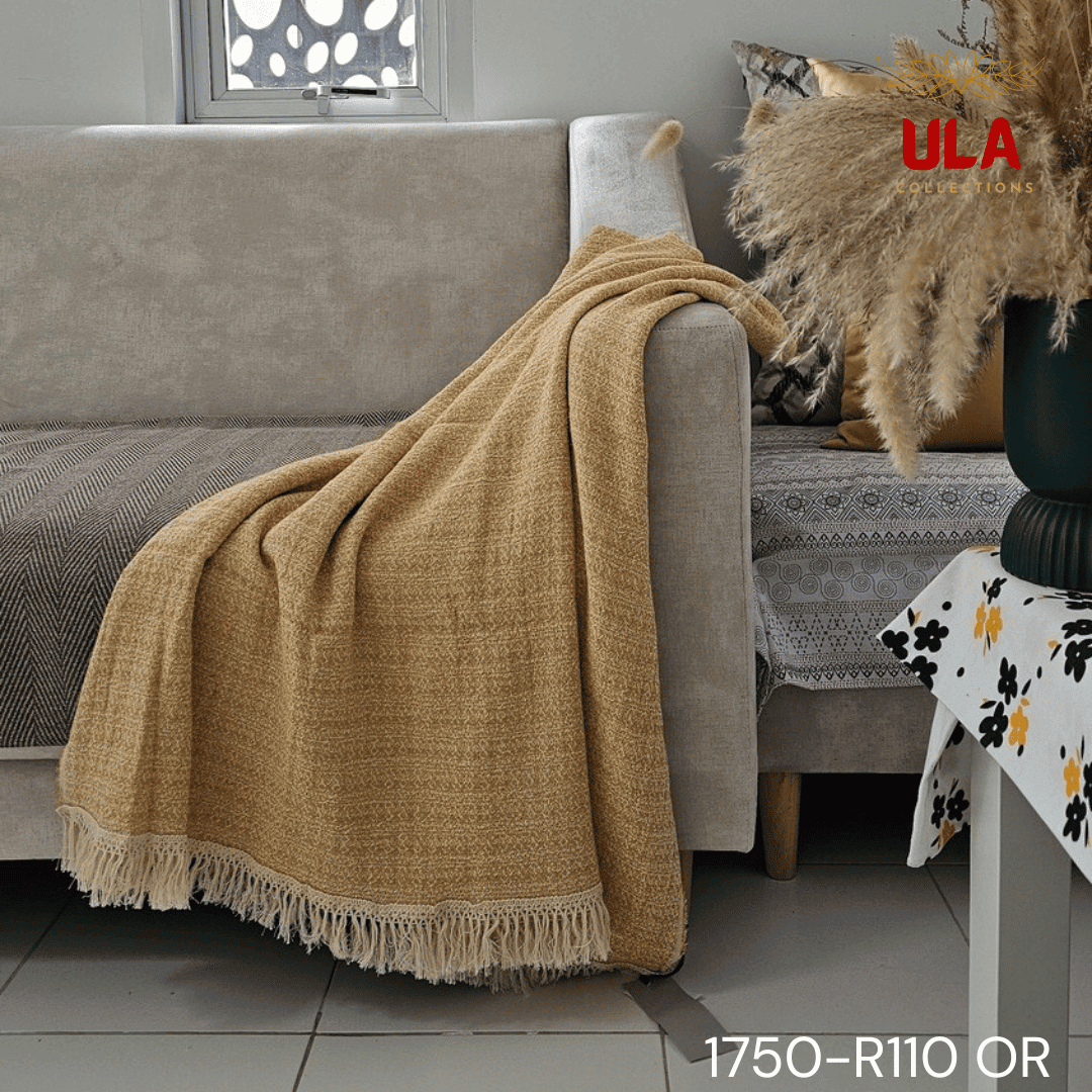 A close-up view of a soft woven throw blanket or textile, primarily in a speckled mustard yellow or marigold color, blended with white threads for a heathered texture. The blanket is finished with a wide white tassel fringe