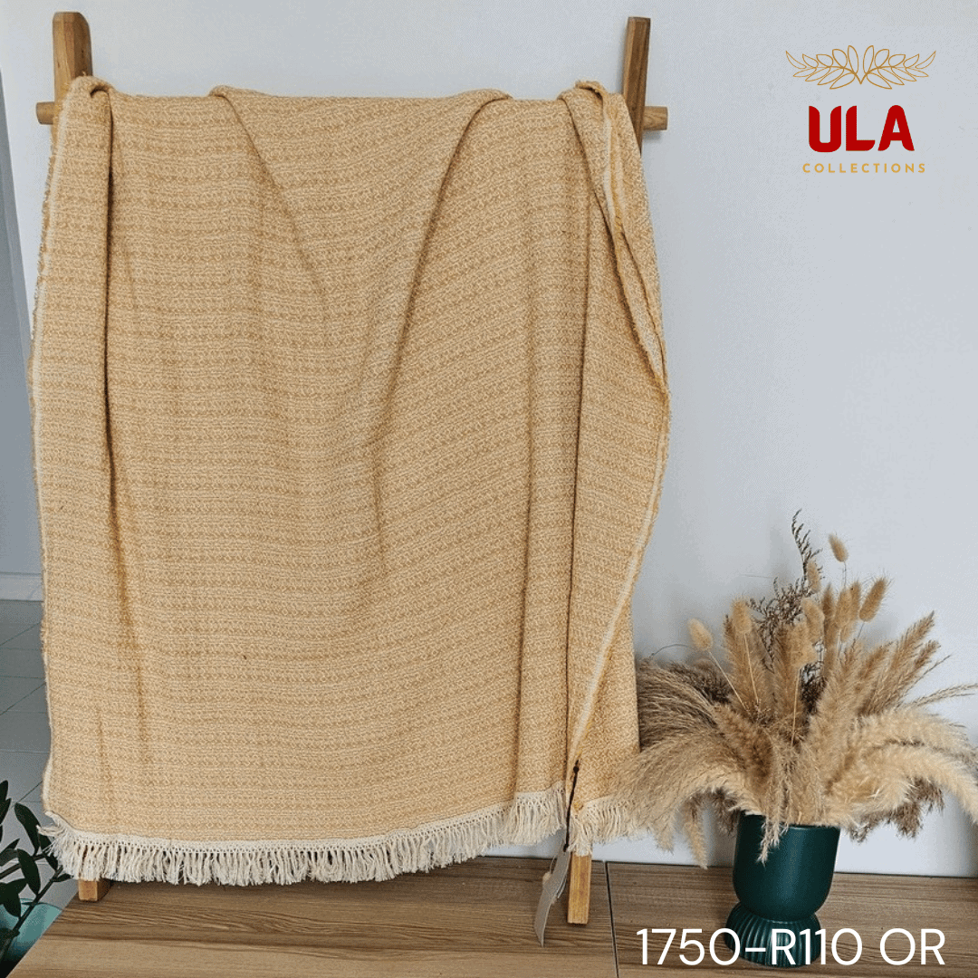 A close-up view of a soft woven throw blanket or textile, primarily in a speckled mustard yellow or marigold color, blended with white threads for a heathered texture. The blanket is finished with a wide white tassel fringe