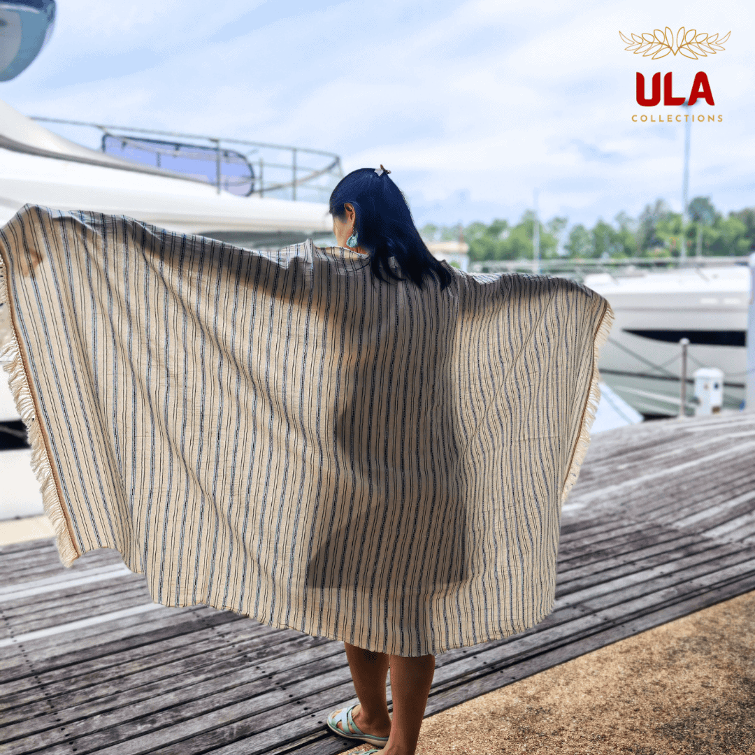 cream throw blanket with thin navy blue and light blue stripes, worn as a shawl by a woman on a wooden dock overlooking the water.
