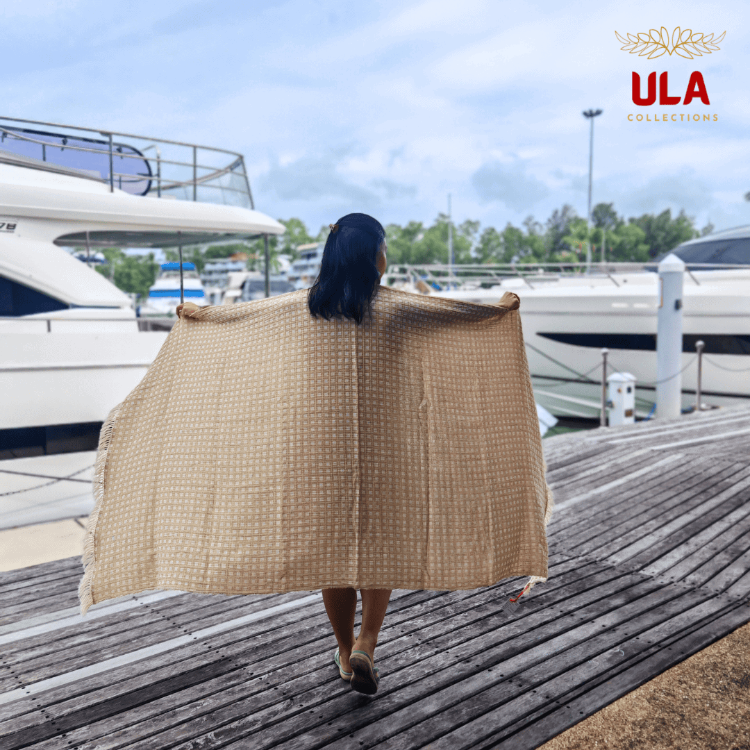 light khaki woven throw blanket with white square check pattern, worn as a shawl by a woman standing on a wooden pier near boats.