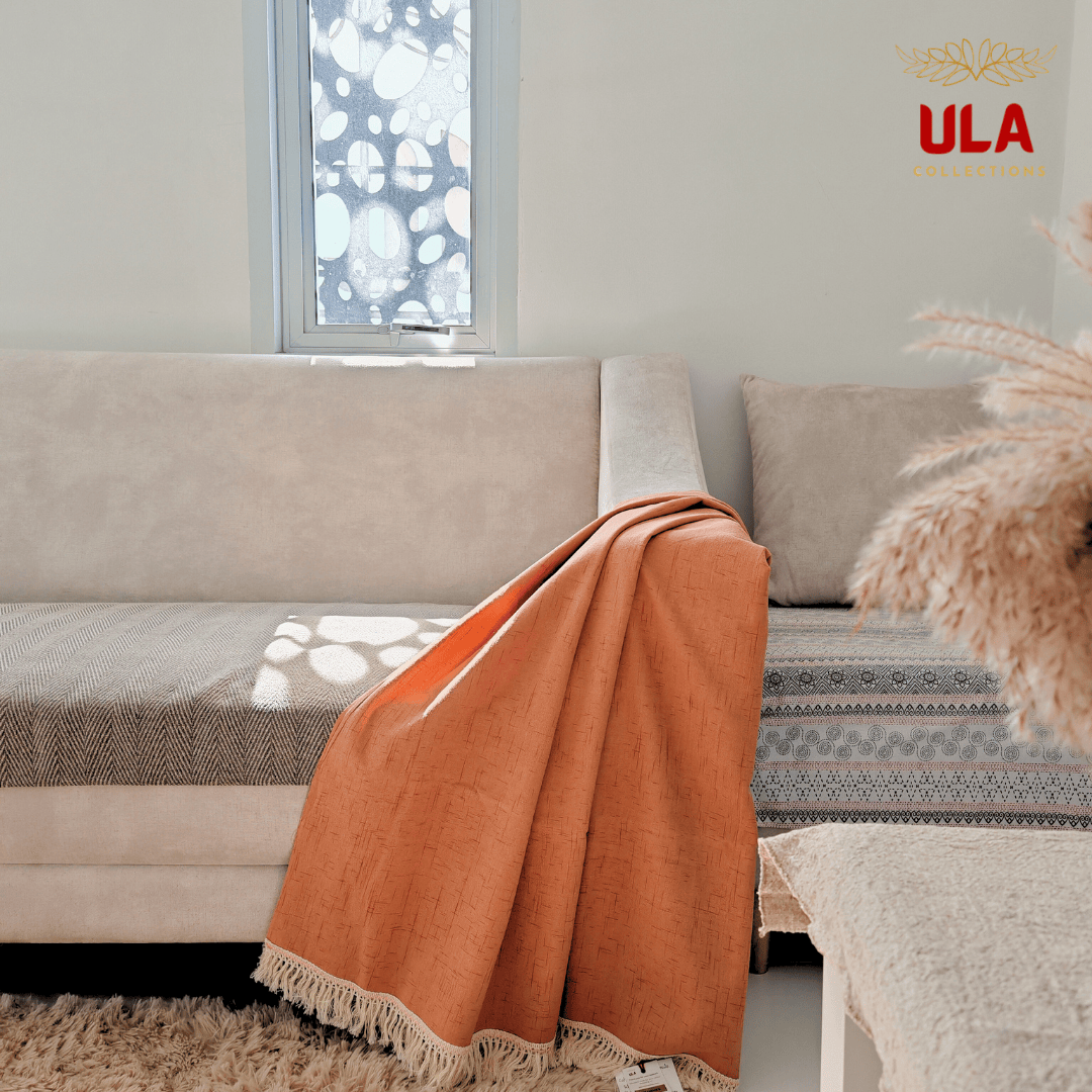 A vibrant terracotta or burnt orange throw blanket with a subtle cross-hatch texture and a cream-colored fringe border is draped over the arm of a beige sofa. The room is decorated in a warm, neutral palette, featuring a plush shaggy rug, a window with decorative frosted glass, and a pampas grass arrangement in the foreground. The "ULA COLLECTIONS" logo is positioned in the upper right corner.