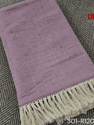 A close-up view of a soft woven throw blanket or textile in a dusky mauve or light purple color. The fabric features a subtle Ikat pattern of short, thin, black dash marks, suggesting a heathered linen-like texture. The blanket is finished with a wide natural beige tassel fringe and a delicate woven trim.