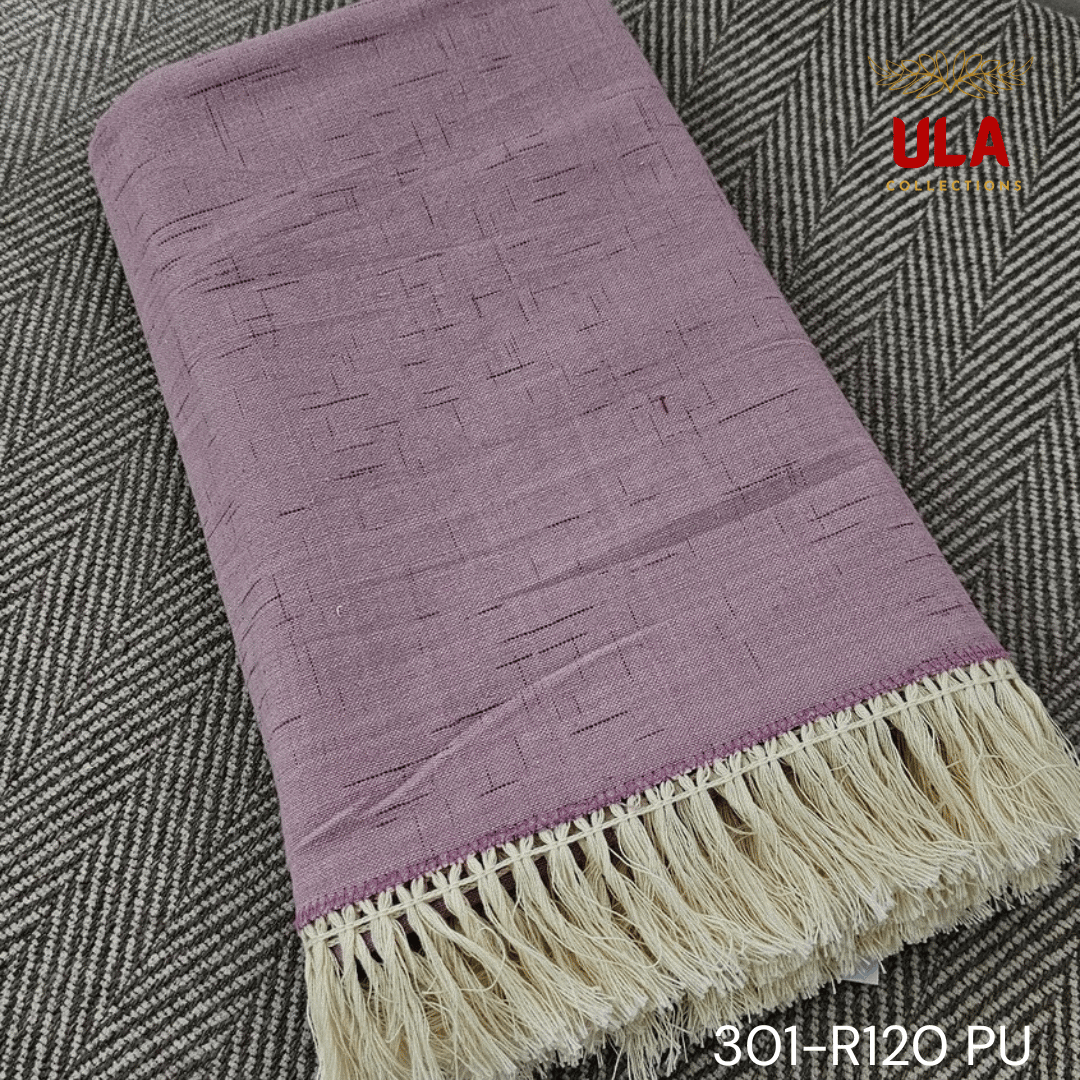 A close-up view of a soft woven throw blanket or textile in a dusky mauve or light purple color. The fabric features a subtle Ikat pattern of short, thin, black dash marks, suggesting a heathered linen-like texture. The blanket is finished with a wide natural beige tassel fringe and a delicate woven trim.
