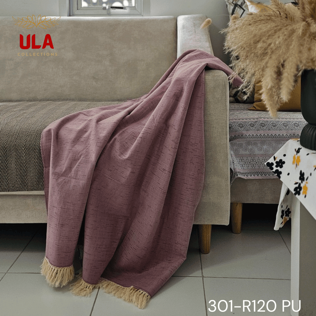 A close-up view of a soft woven throw blanket or textile in a dusky mauve or light purple color. The fabric features a subtle Ikat pattern of short, thin, black dash marks, suggesting a heathered linen-like texture. The blanket is finished with a wide natural beige tassel fringe and a delicate woven trim.