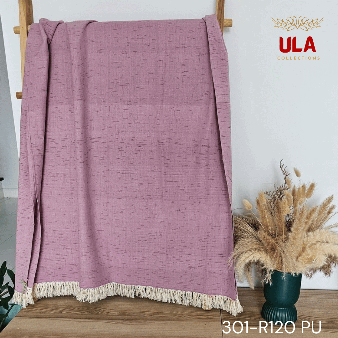 A close-up view of a soft woven throw blanket or textile in a dusky mauve or light purple color. The fabric features a subtle Ikat pattern of short, thin, black dash marks, suggesting a heathered linen-like texture. The blanket is finished with a wide natural beige tassel fringe and a delicate woven trim.