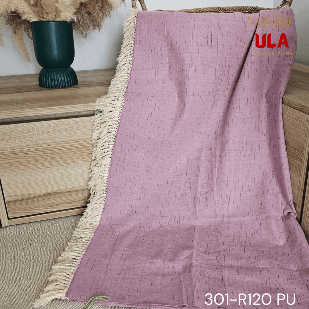 A close-up view of a soft woven throw blanket or textile in a dusky mauve or light purple color. The fabric features a subtle Ikat pattern of short, thin, black dash marks, suggesting a heathered linen-like texture. The blanket is finished with a wide natural beige tassel fringe and a delicate woven trim.