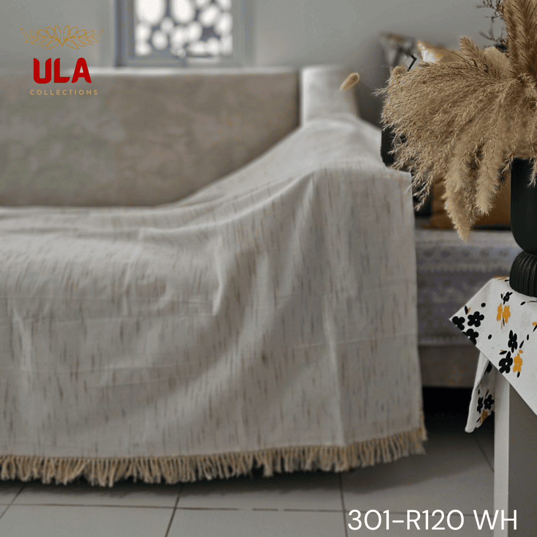 A close-up view of a white woven throw blanket or textile featuring a subtle, vertically dashed Ikat pattern in a thin gold or light brown thread. The blanket is finished with a wide natural beige tassel fringe and is accented with a delicate woven trim.