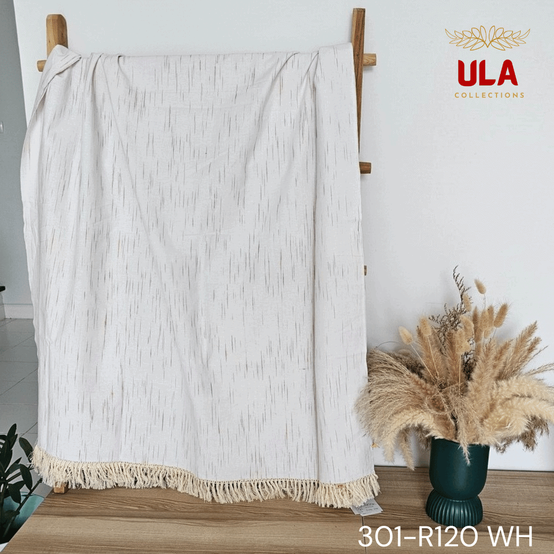 A close-up view of a white woven throw blanket or textile featuring a subtle, vertically dashed Ikat pattern in a thin gold or light brown thread. The blanket is finished with a wide natural beige tassel fringe and is accented with a delicate woven trim.