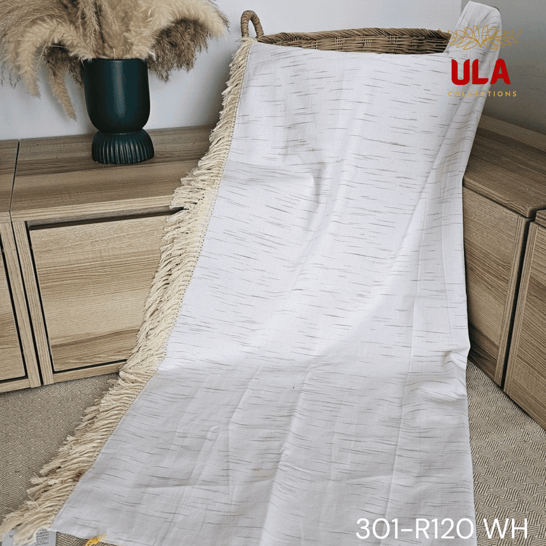 A close-up view of a white woven throw blanket or textile featuring a subtle, vertically dashed Ikat pattern in a thin gold or light brown thread. The blanket is finished with a wide natural beige tassel fringe and is accented with a delicate woven trim.