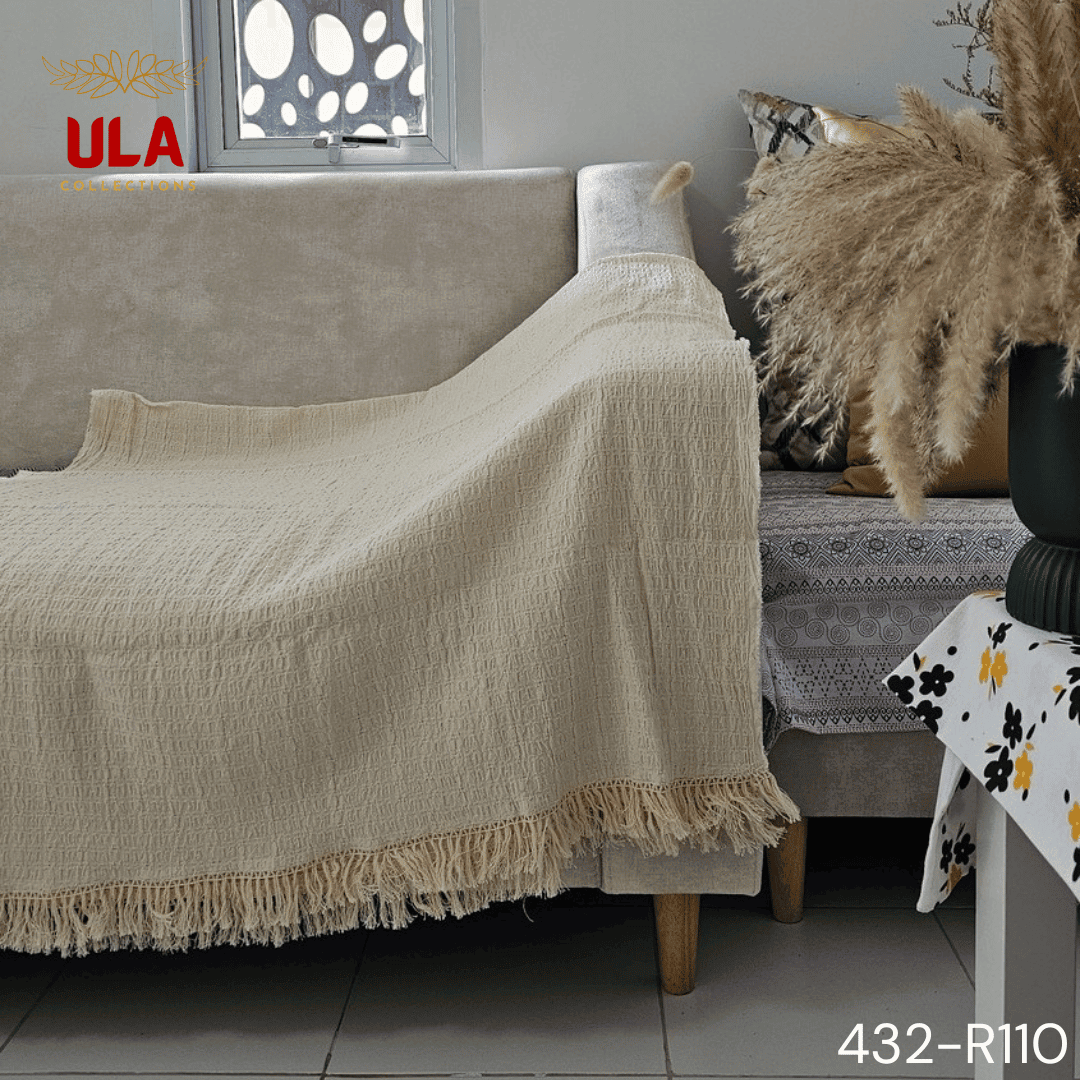 A folded cream or off-white woven throw blanket with a subtle plaid or windowpane pattern in its weave. The textile is finished with a wide natural-colored tassel fringe