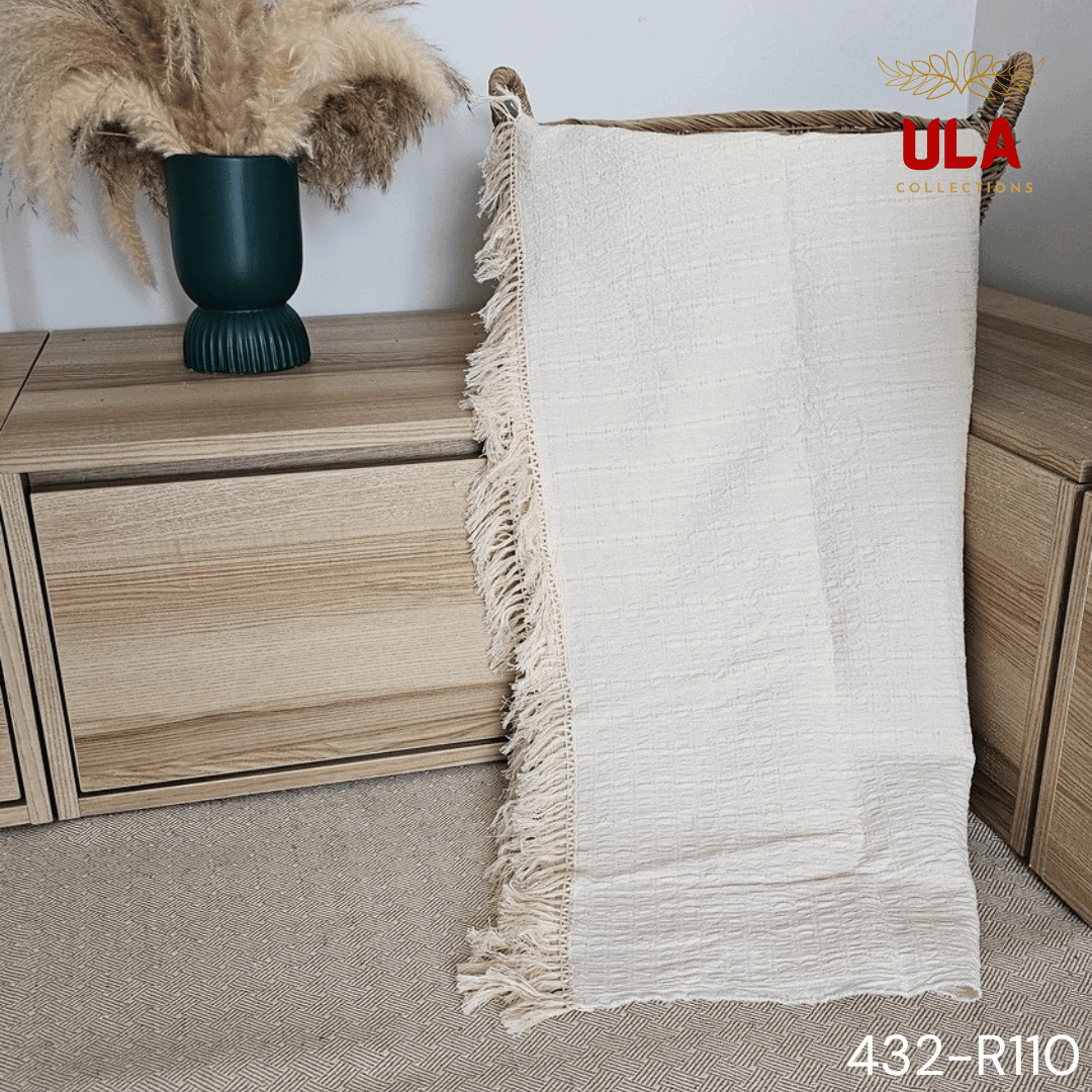 A folded cream or off-white woven throw blanket with a subtle plaid or windowpane pattern in its weave. The textile is finished with a wide natural-colored tassel fringe