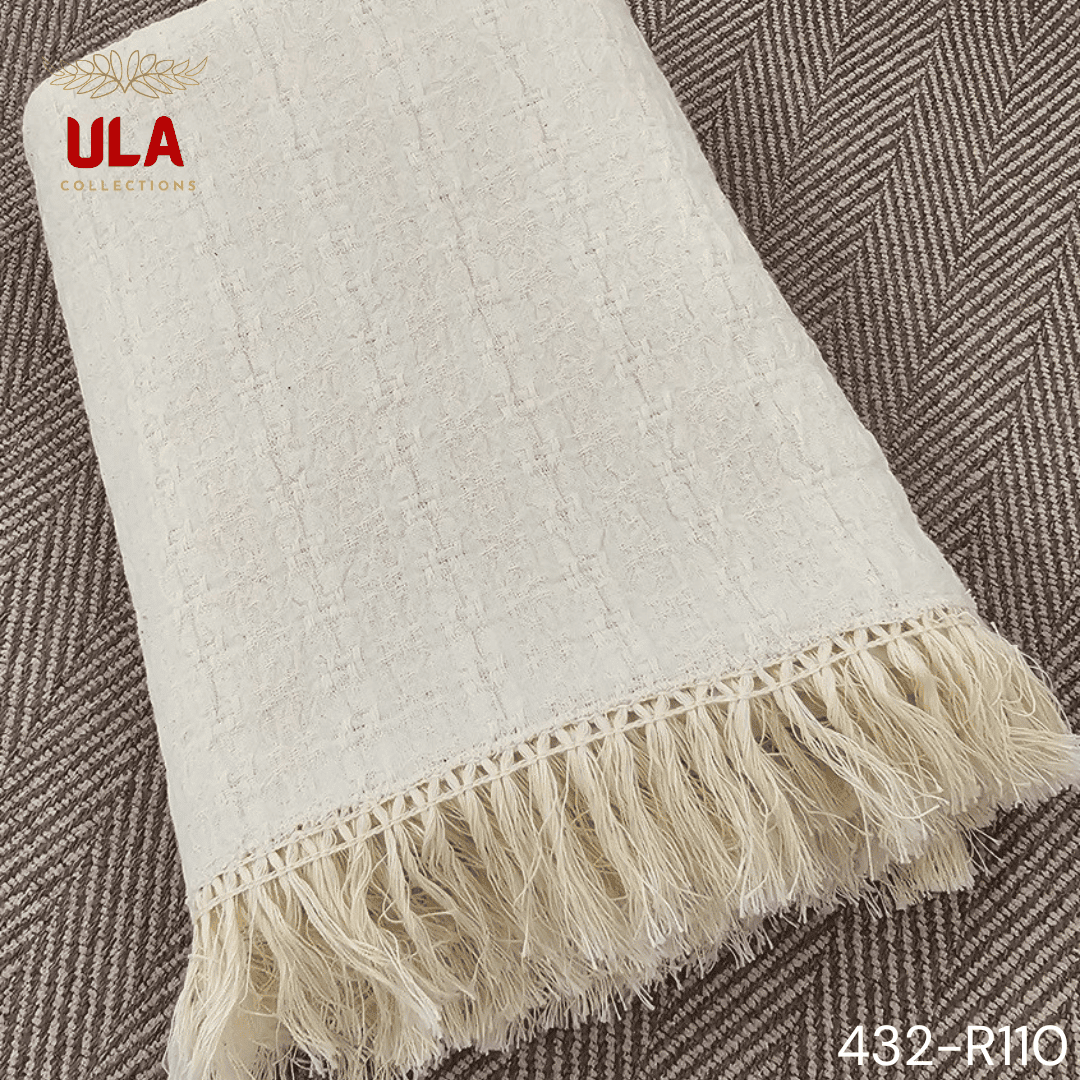 A folded cream or off-white woven throw blanket with a subtle plaid or windowpane pattern in its weave. The textile is finished with a wide natural-colored tassel fringe