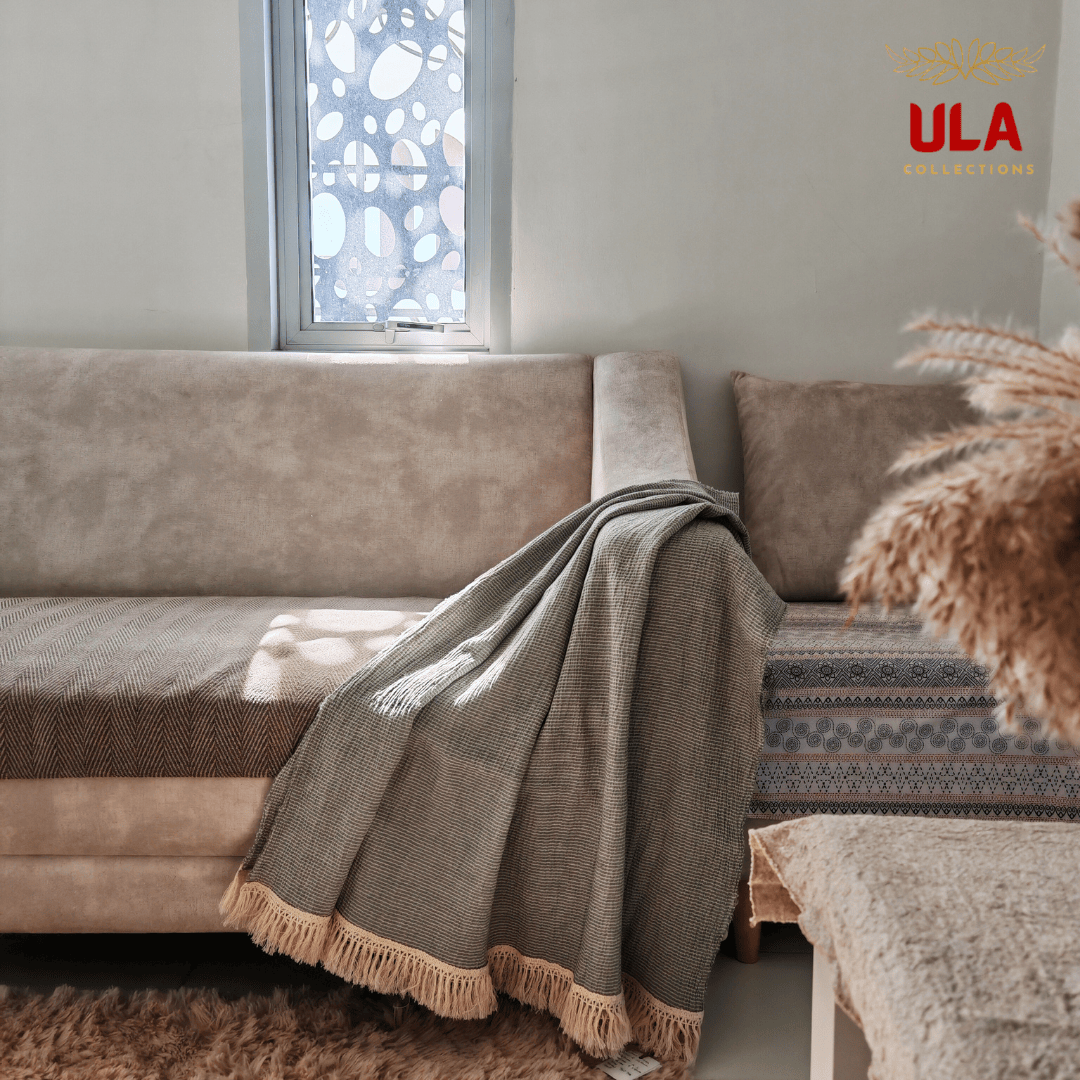 A sage green, textured throw blanket with a thick, cream-colored fringe border draped diagonally over the arm and seat of a beige sofa. The room is styled with neutral tones, featuring a fluffy brown rug and a window with decorative frosted glass in the background. A "ULA Collections" logo is visible in the top right corner.
