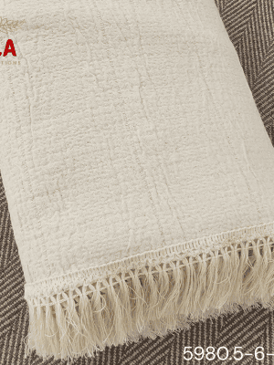 A folded cream or off-white woven throw blanket with a prominent basketweave or waffle knit texture. The textile is finished with a wide natural-colored tassel fringe and is displayed draped over a couch or background fabric with a dark gray herringbone pattern, highlighting a modern farmhouse or cozy minimalist home decor style.