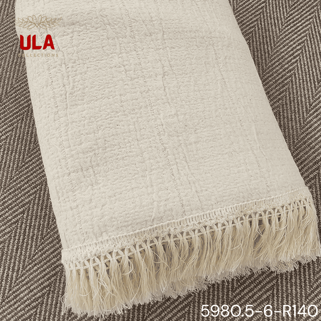 A folded cream or off-white woven throw blanket with a prominent basketweave or waffle knit texture. The textile is finished with a wide natural-colored tassel fringe and is displayed draped over a couch or background fabric with a dark gray herringbone pattern, highlighting a modern farmhouse or cozy minimalist home decor style.