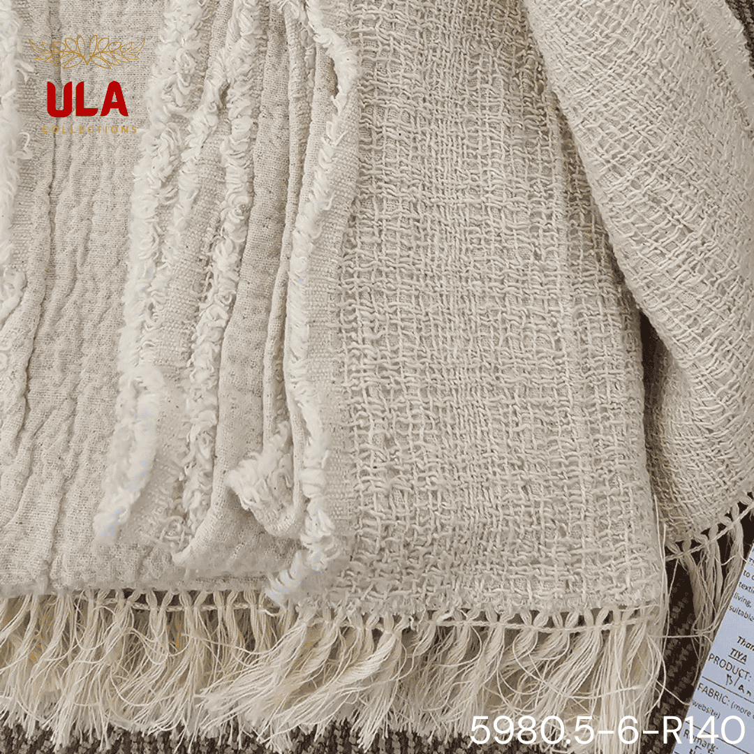 A folded cream or off-white woven throw blanket with a prominent basketweave or waffle knit texture. The textile is finished with a wide natural-colored tassel fringe and is displayed draped over a couch or background fabric with a dark gray herringbone pattern, highlighting a modern farmhouse or cozy minimalist home decor style.