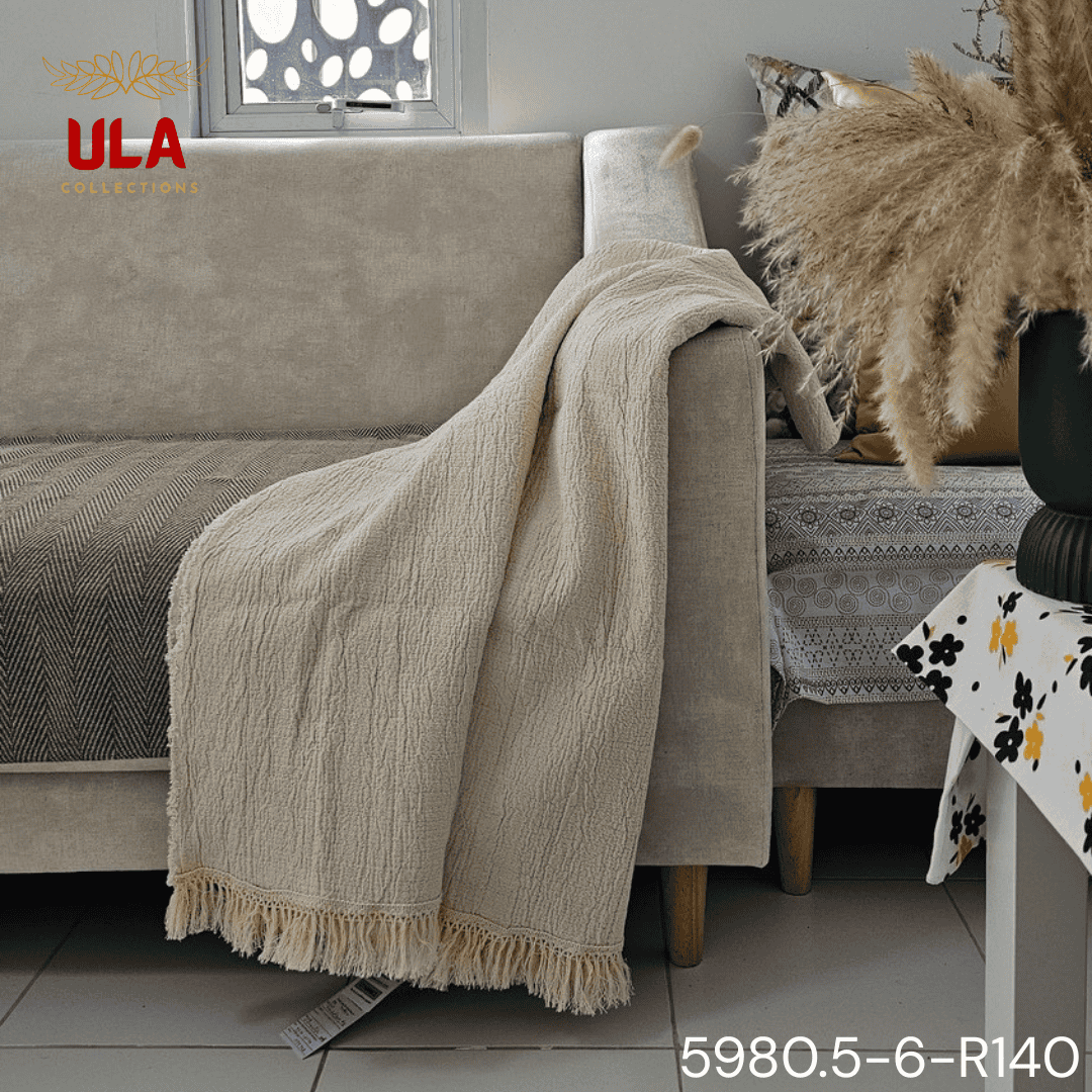 A folded cream or off-white woven throw blanket with a prominent basketweave or waffle knit texture. The textile is finished with a wide natural-colored tassel fringe and is displayed draped over a couch or background fabric with a dark gray herringbone pattern, highlighting a modern farmhouse or cozy minimalist home decor style.