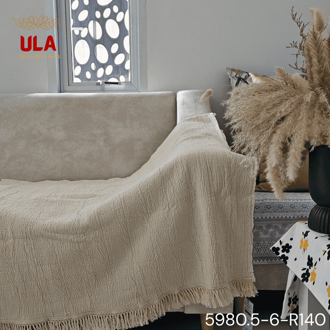 A folded cream or off-white woven throw blanket with a prominent basketweave or waffle knit texture. The textile is finished with a wide natural-colored tassel fringe and is displayed draped over a couch or background fabric with a dark gray herringbone pattern, highlighting a modern farmhouse or cozy minimalist home decor style.