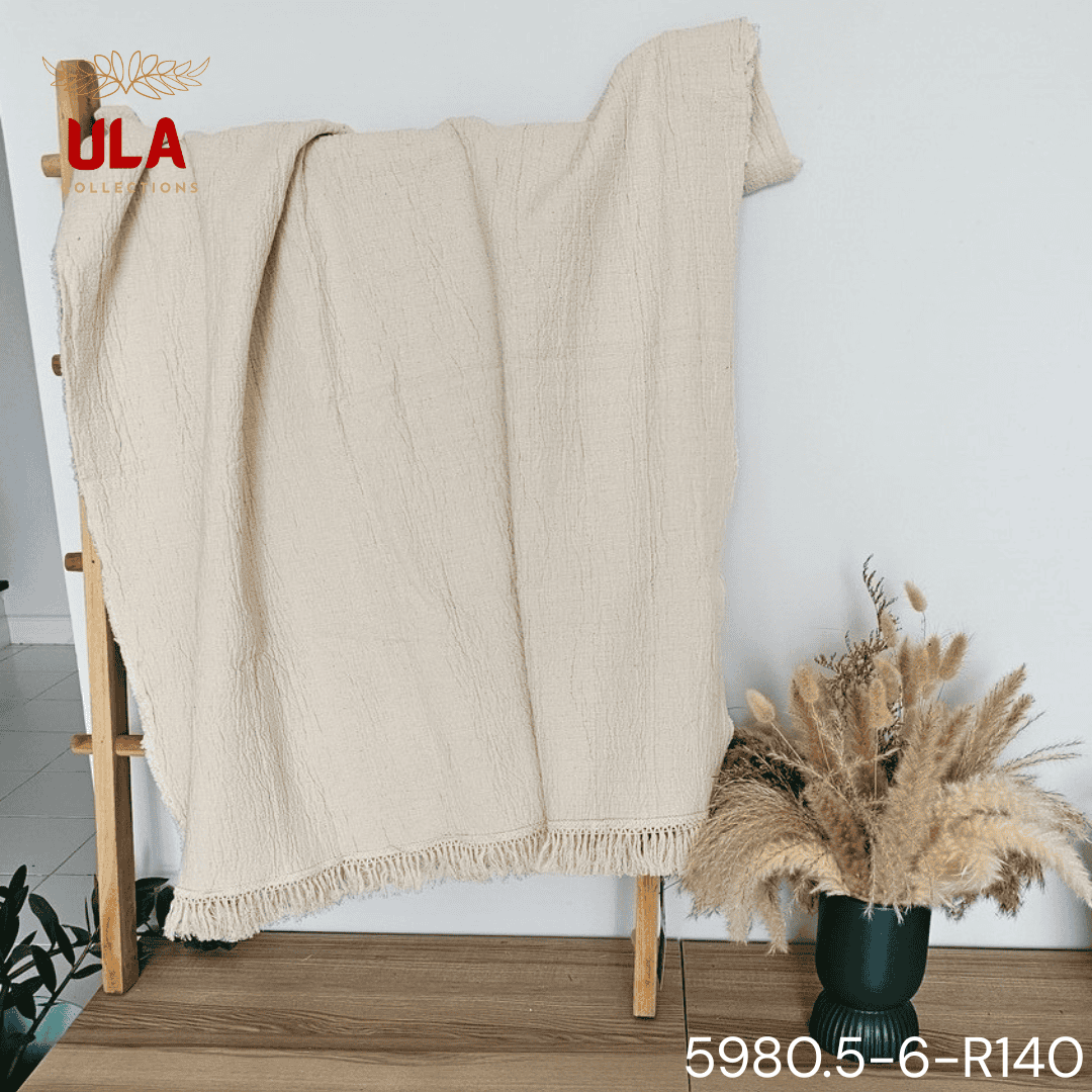 A folded cream or off-white woven throw blanket with a prominent basketweave or waffle knit texture. The textile is finished with a wide natural-colored tassel fringe and is displayed draped over a couch or background fabric with a dark gray herringbone pattern, highlighting a modern farmhouse or cozy minimalist home decor style.