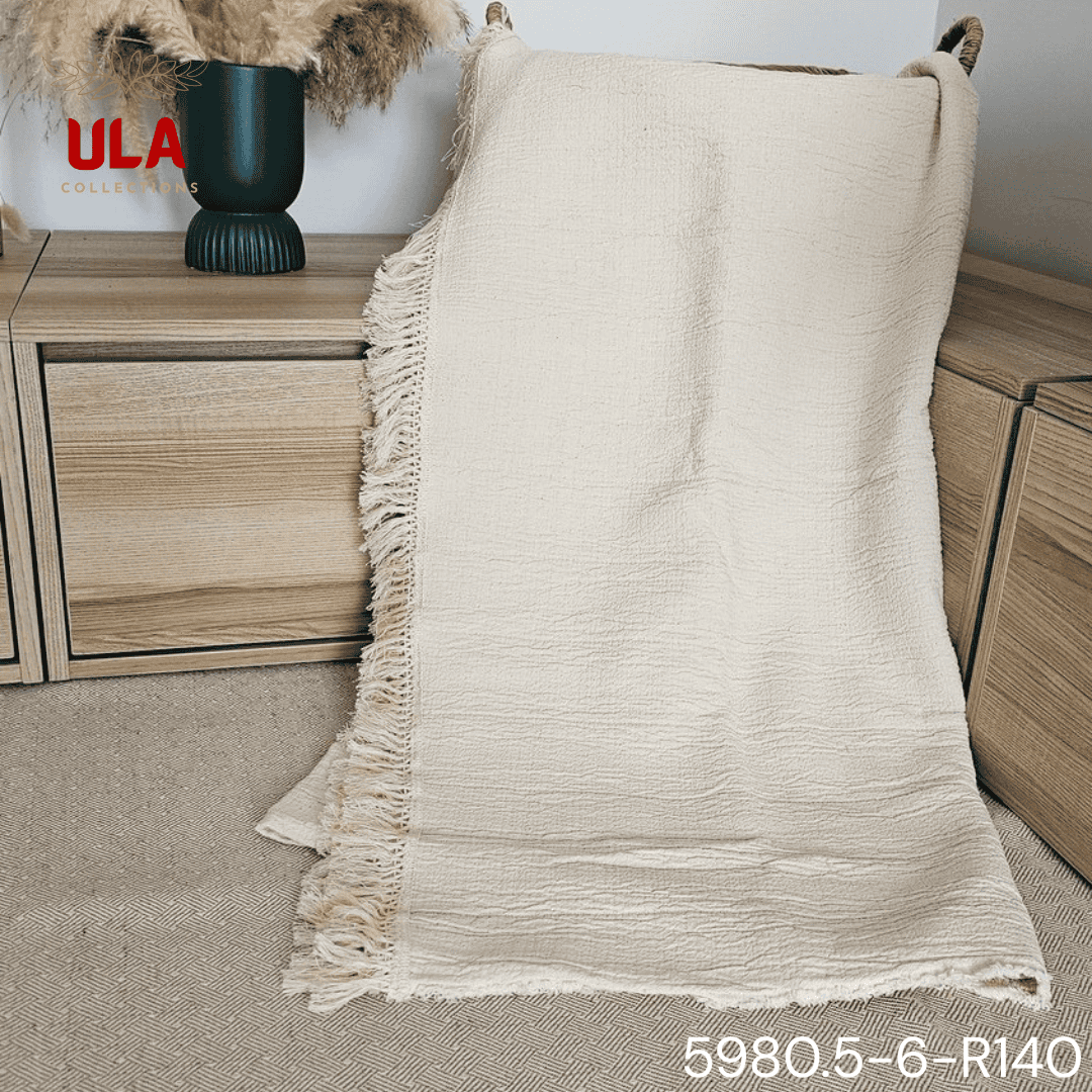A folded cream or off-white woven throw blanket with a prominent basketweave or waffle knit texture. The textile is finished with a wide natural-colored tassel fringe and is displayed draped over a couch or background fabric with a dark gray herringbone pattern, highlighting a modern farmhouse or cozy minimalist home decor style.