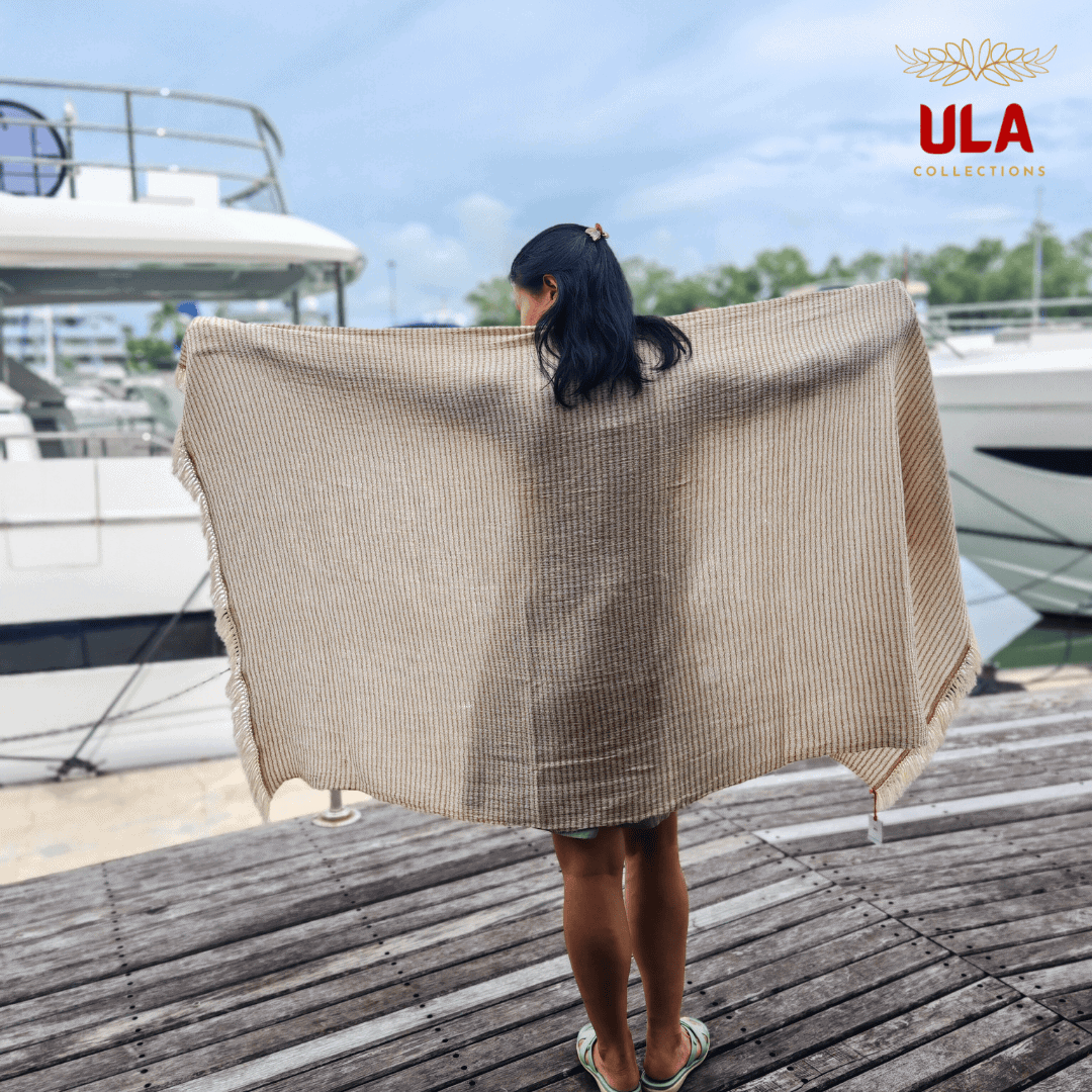 striped woven throw blanket in tan/beige with thin green stripes, draped over a woman standing backward on a wooden marina dock with luxury yachts.