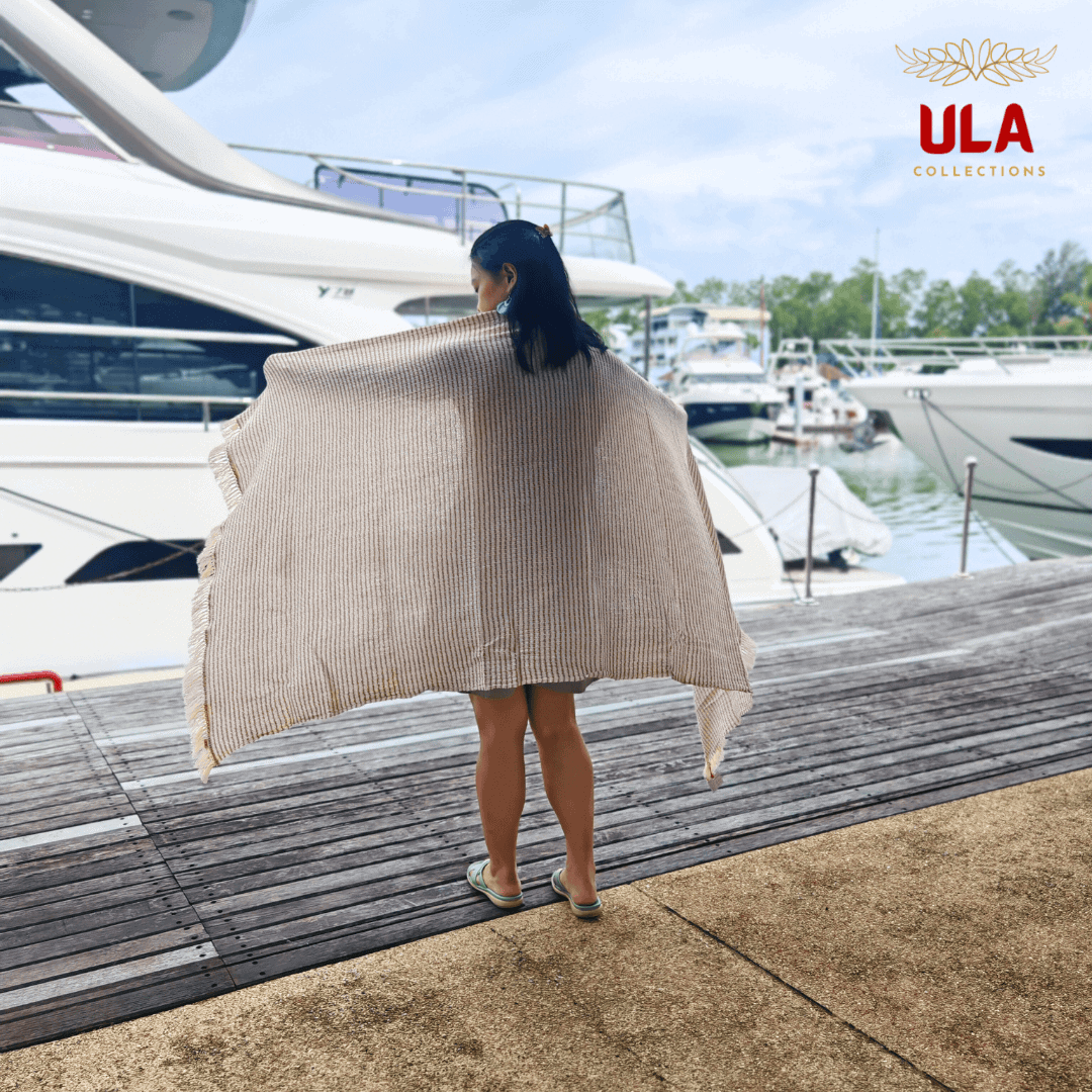 striped woven throw blanket in cream/tan with thin red-brown stripes, worn as a shawl by a woman standing on a wooden pier near yachts.