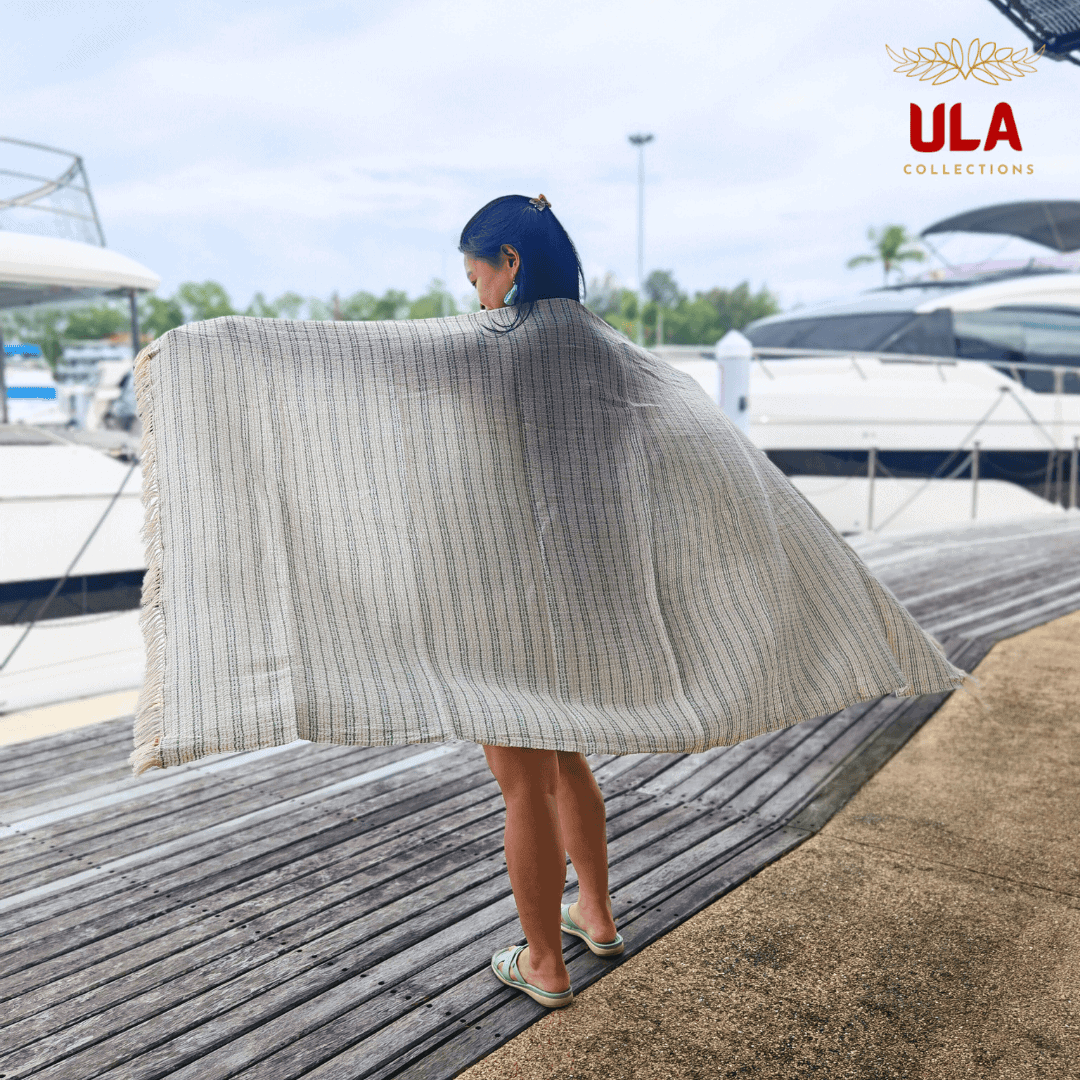 striped woven throw blanket in cream with thin sage green stripes, worn as a shawl by a woman standing on a wooden dock with boats visible behind her.